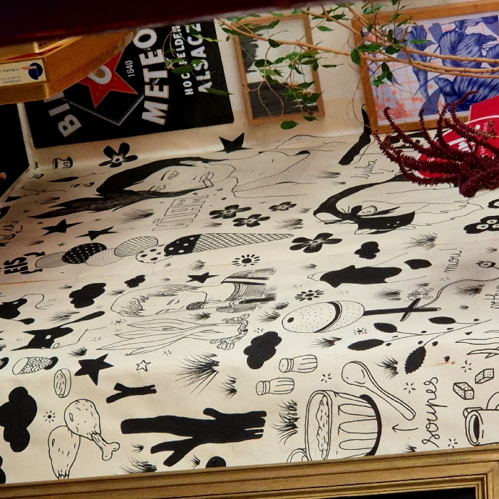 The image depicts an interior corner of a cafe or restaurant located in Strasbourg, France. The dominant feature is a wall extensively decorated with hand-drawn, black-on-white illustrations in a whimsical, cartoonish style. These illustrations include food items such as chicken drumsticks, burgers, ice cream cones (one labeled "EIS"), salt and pepper shakers, a bowl of soup labeled "soupes" with an accompanying spoon, and a coffee cup with sugar cubes. Natural elements like trees, grass tufts, clouds, stars, and flowers are interspersed. The wall also features stylized human faces or busts, a dog-like figure, and handwritten words including "VIERA," "ljuba," "miau!", and "arthur...". To the upper right, a black poster for "METEOR" beer is visible, displaying "1640," "HOCHFELDEN," and "ALSACZ," indicating the brewery's origin in Alsace. Below this poster, a small, simple wooden-framed picture and a larger wooden-framed print with abstract blue and white designs are mounted. A vibrant red, feathery plant, possibly Amaranthus, and a green-leaved plant with thin brown stems are positioned in the foreground, partially obscuring the lower right wall and artwork. A portion of a golden-colored frame is visible on the far-left edge of the image. No individuals are present in the scene.