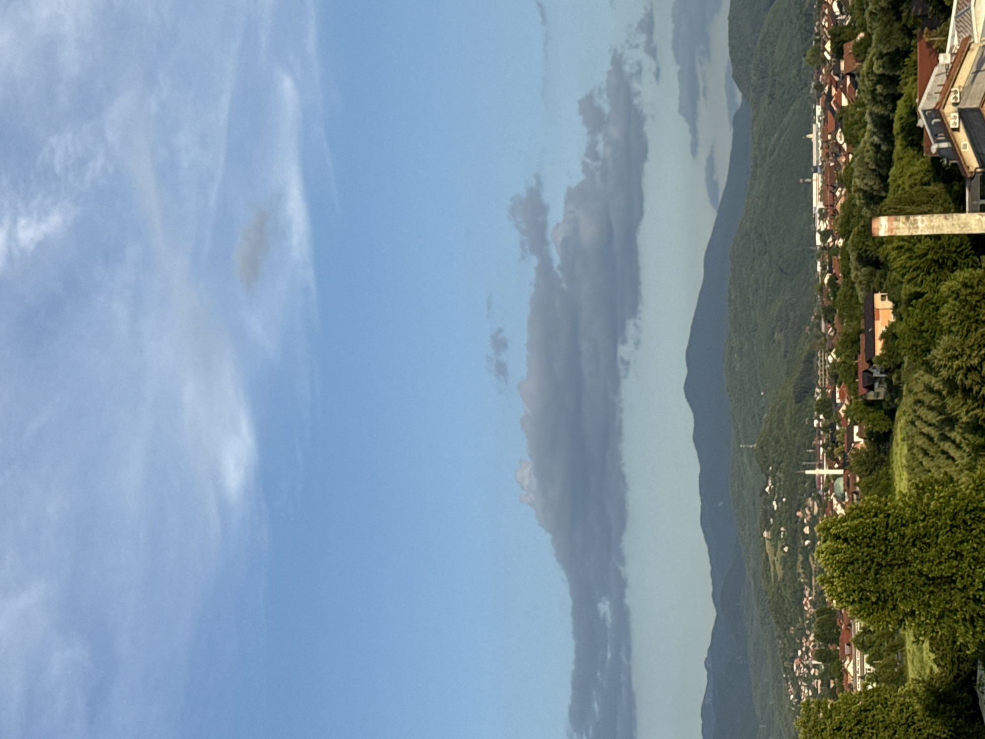 An aerial view of the city of Ilidža, Bosnia and Herzegovina, captures a sprawling landscape under a partly cloudy sky. In the foreground, lush green trees dominate, with a few buildings peeking through the foliage. The middle ground reveals a dense urban area with red-tiled roofs, interspersed with more trees and a prominent mosque with a white minaret. Further in the background, rolling green hills rise, culminating in a range of mountains that fade into the distance. The sky is a soft blue, with scattered clouds, some appearing dark and heavy, suggesting the possibility of rain. No people are visible in the image.