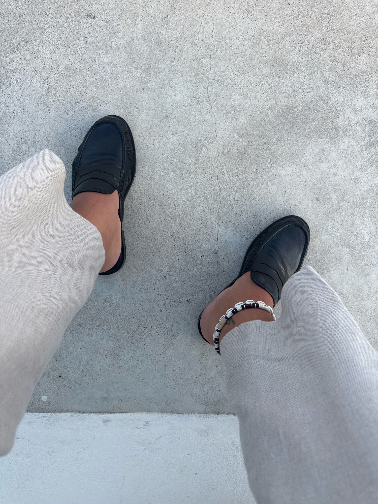 This is a top-down, full-body shot of a person wearing light-colored linen pants and black leather mules. The person's left leg is on the left side of the frame, and their right leg is on the right side. Both feet are clad in black, backless leather loafers with a penny loafer detail across the vamp. The person is wearing a beaded anklet on their right ankle, featuring black, white, and tan beads. The background is a rough, light grey concrete surface, with a white-painted step or ledge at the bottom of the frame. The scene suggests an outdoor setting, possibly a courtyard or public space within Mafra, Portugal.