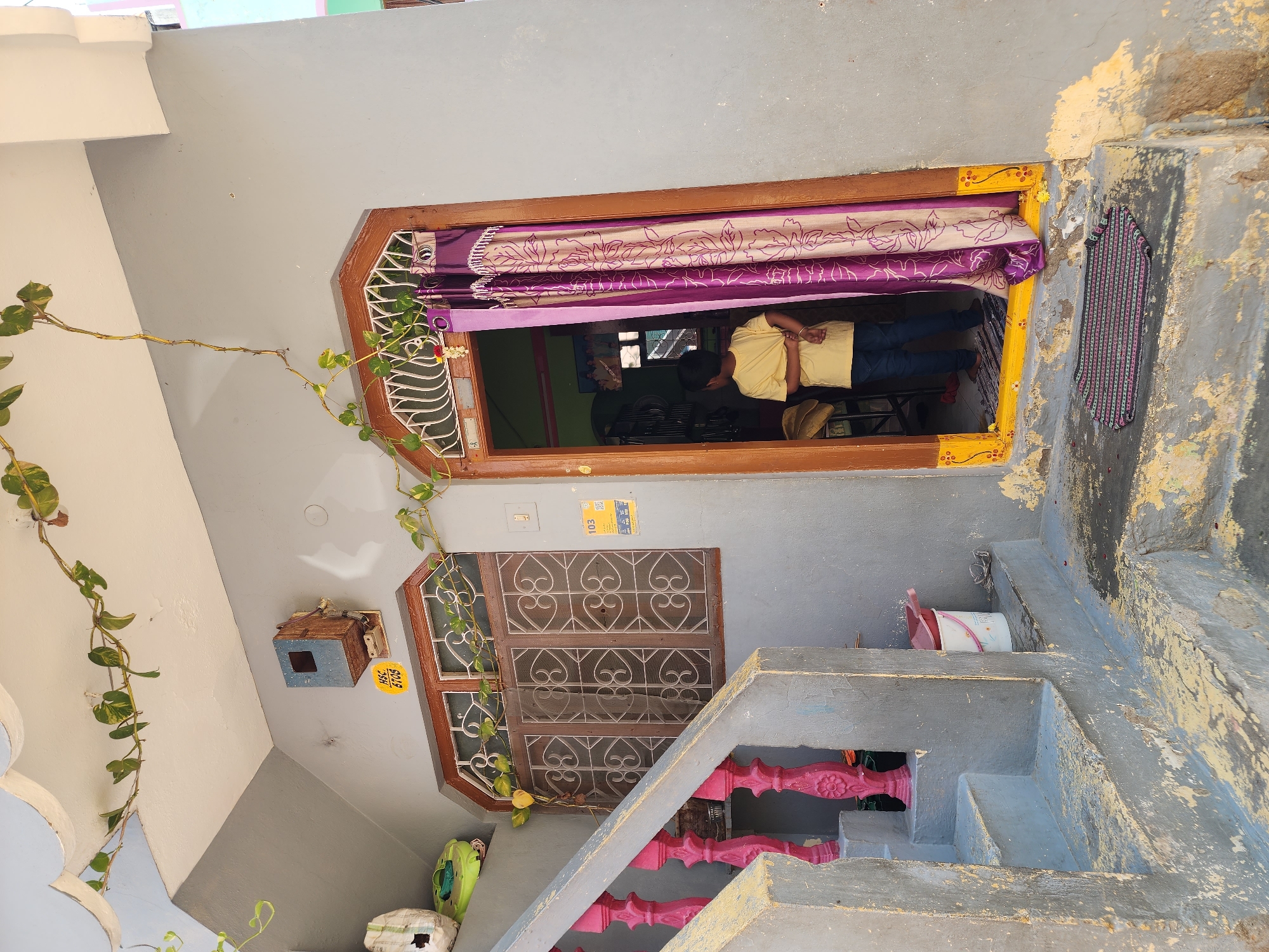 The image depicts the entrance to a residence in Pedda Kammavaripalli, India. A young child, dressed in a yellow t-shirt and dark trousers, stands facing inwards within the open doorway, positioned between a patterned purple and beige curtain on the right and the interior of the home on the left. The doorway features a dark brown wooden frame with a vibrant yellow trim adorned with painted floral motifs along the bottom. A dark grey and red striped door mat lies on the worn concrete step directly outside the doorway.
To the left of the doorway, a set of concrete steps leads upwards, featuring a balustrade with prominent pink, ornate balusters and a grey handrail. The walls of the structure are painted a light grey, showing signs of wear and peeling paint, especially on the steps and lower sections. Above the steps and to the left of the door, a rectangular window with a dark brown wooden frame is fitted with white, intricate metal grilles. Above this window, a utility box, possibly an electrical meter or junction box, is mounted on the wall. A yellow sticker with "HSC 6708" is affixed below it. Further to the right of the window, near the door frame, a white light switch is visible, along with a yellow sticker displaying "103" and a QR code below it.
Overhead, a light-colored soffit or overhang is adorned with a trailing green vine with yellow-green leaves. Another section of the vine extends downwards near the window. On the upper landing, partially visible behind the balustrade, a white plastic bucket with a red handle and a green object are present. A white bucket with a pink lid is positioned under the upper part of the stairs. The overall scene suggests a lived-in residential environment with a mix of functional utility and decorative elements.