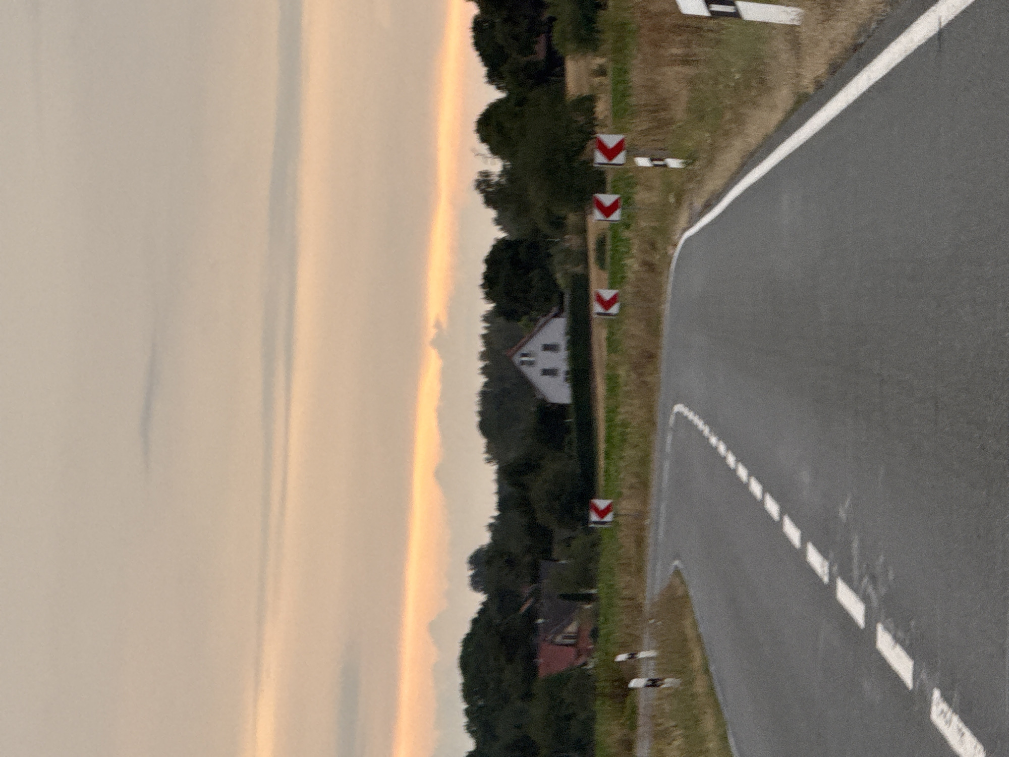 The image captures a rural road in Hohenstein, Germany, at dusk. The road, paved with asphalt, curves to the right. A dashed white line marks the center of the road, with solid white lines delineating the edges. Along the side of the road, several white posts with red and white directional arrows indicate a curve. In the background, a small cluster of trees frames a rural landscape featuring a few houses with distinctive red roofs and a larger, lighter-colored house with a prominent gable. Beyond the houses, a dense line of trees creates a dark silhouette against a sky illuminated by the warm hues of the setting sun. Faint mountain ranges are visible in the far distance, hazy against the colorful sky. The environment appears dry, with patches of tall, golden-brown grass visible on the roadside. The overall scene suggests a quiet, rural setting during the twilight hours.