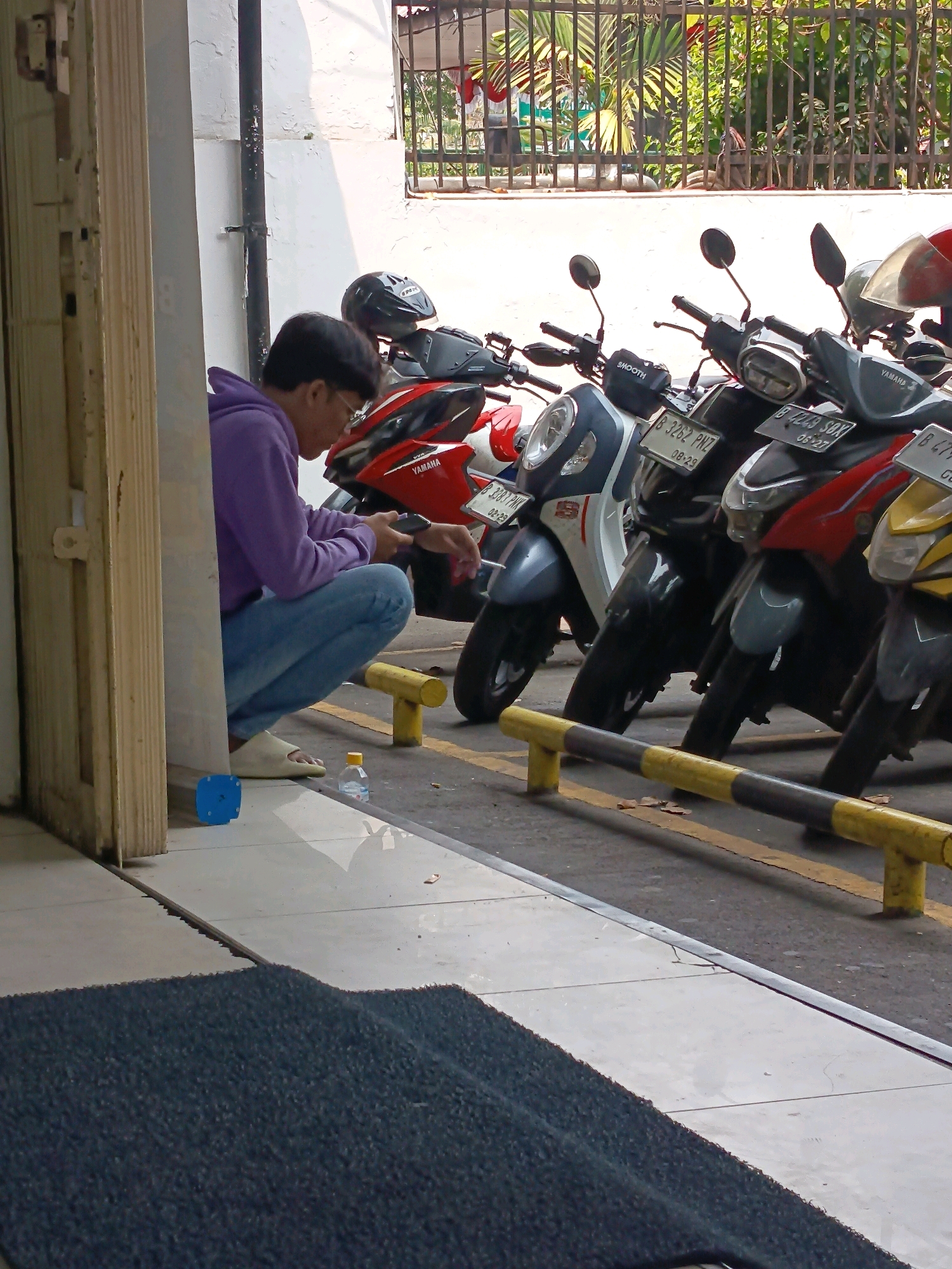 A person wearing a purple hoodie and blue jeans is squatting on the ground next to a row of parked motorcycles. They are looking down at a black object in their hands, possibly a smartphone. A black helmet rests on the seat of one of the motorcycles. In the foreground, a dark blue mat is visible, and a small clear plastic bottle of water sits on the tiled ground. The background shows a black metal fence with foliage behind it, suggesting an outdoor area. The scene appears to be a parking lot, likely in a commercial or residential area of Central Jakarta, Indonesia.
