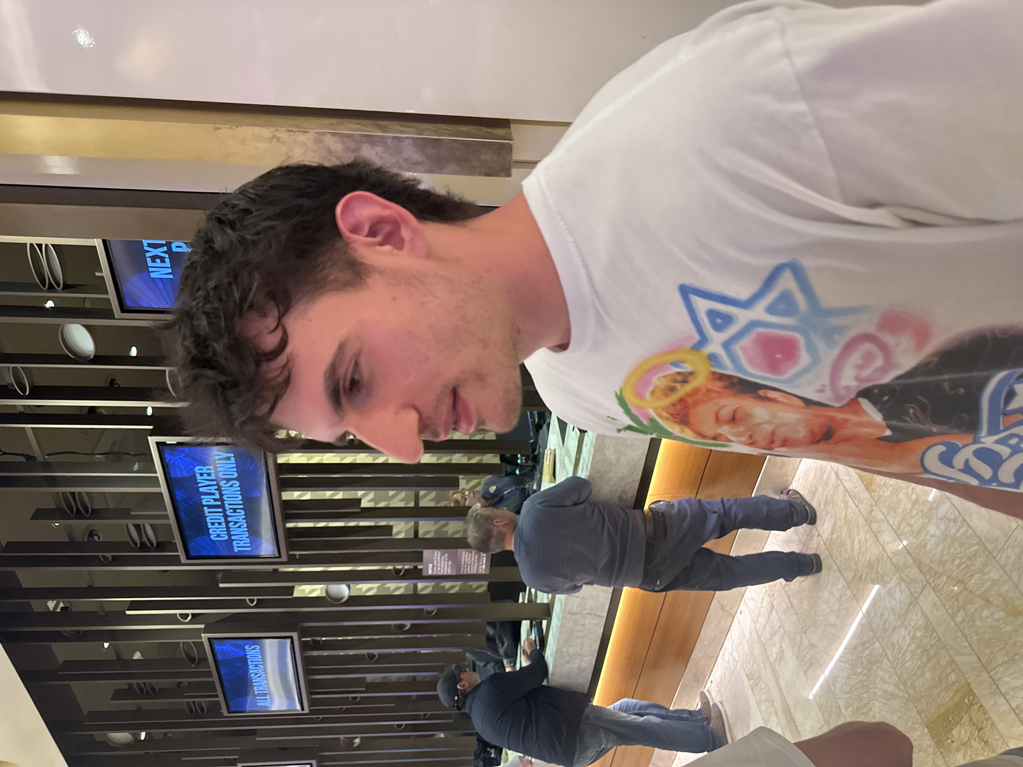 The photograph shows the interior of what appears to be a casino or financial transaction center. The foreground is dominated by the face of a young man in a white T-shirt with colorful graphics. Behind him, a queue of people waits at the counter. Electronic displays above the counter are visible, displaying text such as "CREDIT PLAYER TRANSACTIONS ONLY" and "ALL TRANSACTIONS." The floor is tiled with a light-colored, marbled pattern. The photograph captures a casual, candid moment with a focus on the subject in the foreground and the backdrop of a service-oriented location. While the city cannot be definitively determined from the visual elements alone, the prompt specifies the setting as Fort Lauderdale, United States of America.
