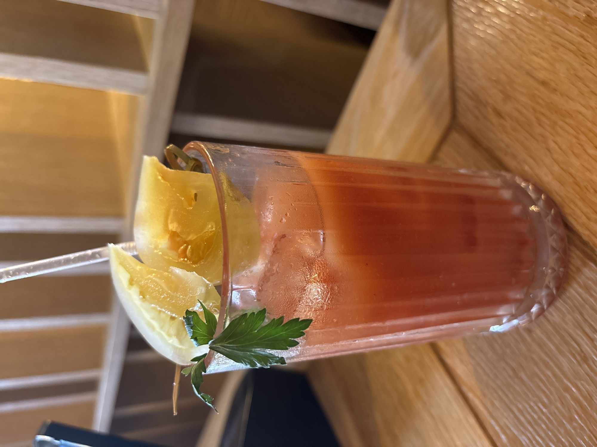 A tall glass of red liquid, possibly a cocktail or juice, sits on a wooden table. The glass is fluted and filled with ice and the drink, which is garnished with a sprig of parsley and two slices of what appear to be citrus fruit and pineapple. A clear plastic straw is also visible in the glass. The background is blurred but suggests an indoor setting with wooden shelves and warm lighting. The image does not provide any information to indicate a location in Saint Petersburg, Russia.