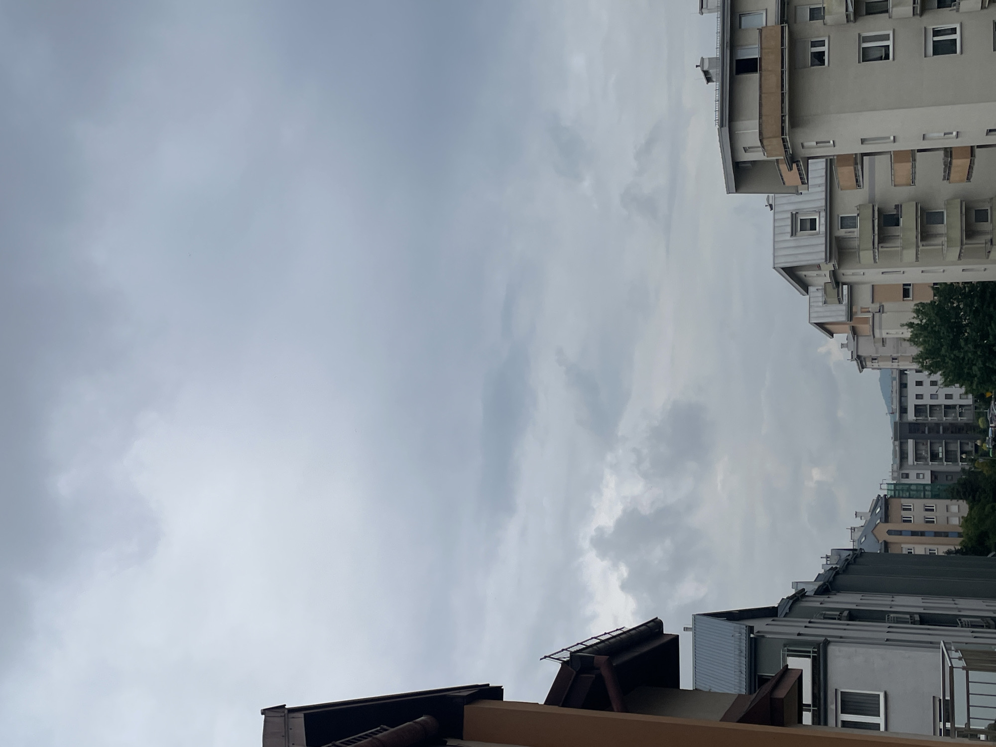The image depicts a view of a cloudy sky framed by buildings in an urban environment. The buildings on the right side of the image are taller, likely apartment buildings, with visible windows and balconies. A portion of a green tree is also visible. The buildings on the left are shorter, with visible rooftops and architectural details. The sky is predominantly gray, indicating overcast weather. The image is rotated, with the sky appearing "sideways" between the building structures.
