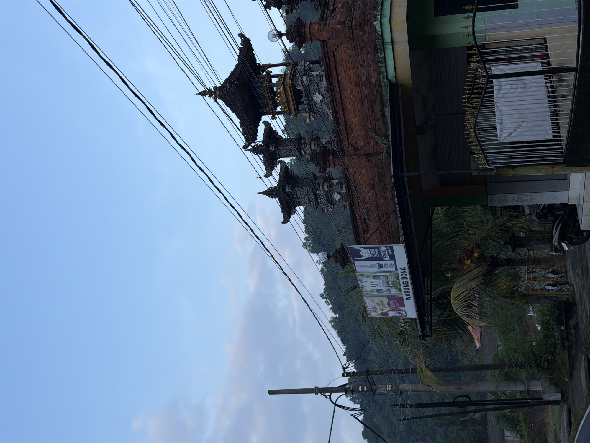 A street-level outdoor view captures a building with traditional Balinese architectural elements in Manggis, Indonesia, during twilight. The sky is a soft blue with wispy clouds, and a dense network of electrical wires crisscrosses the upper portion of the frame.
The building features a red brick facade and a tiered, dark-tiled roof. Ornate stone statues with tiered roofs sit atop the building, resembling miniature temples. A sign for "WARUNG DONA" is visible, along with several advertisements for beverages. In front of the building, a palm tree laden with coconuts stands tall. A metal fence with decorative finials encloses a portion of the property. A scooter is parked near the fence. The background consists of lush green hills and vegetation, suggesting a rural or semi-rural setting. A utility pole with multiple cables is prominent on the left side of the image. The overall lighting suggests either early morning or late evening.