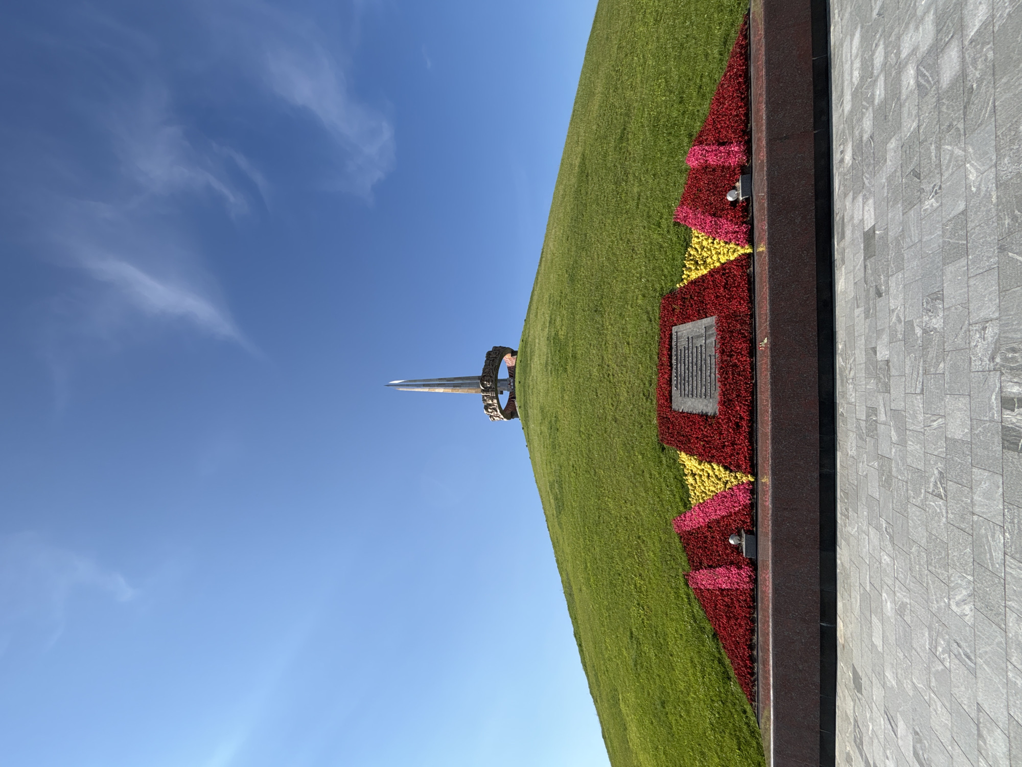 This is a wide, low-angle, full-shot photograph of the **Kurgan of Glory (Mound of Glory)** in the Ozyaritska-Slabadski rural council, Belarus. The monument is situated on a large, grassy mound, which takes up the majority of the frame. Atop the mound, a tall, slender obelisk culminates in a sharp point, surrounded by a circular structure adorned with reliefs. A ring of golden embellishments is visible around the base of the obelisk.
In front of the mound, a rectangular base of dark granite supports a vibrant flowerbed. The flowerbed is arranged in a dynamic pattern of red and yellow blossoms, creating a stark contrast against the green grass and blue sky. A dark stone plaque with inscribed text is centered within the flowerbed. The foreground consists of a paved area with large, grey stone tiles, arranged in a regular pattern.
The sky above is a clear, bright blue with a few wispy white clouds scattered across the upper portion of the frame. The lighting suggests it is a sunny day. There are no people visible in the image. The scene is immaculately maintained, with the grass neatly cut and the flowers well-tended.