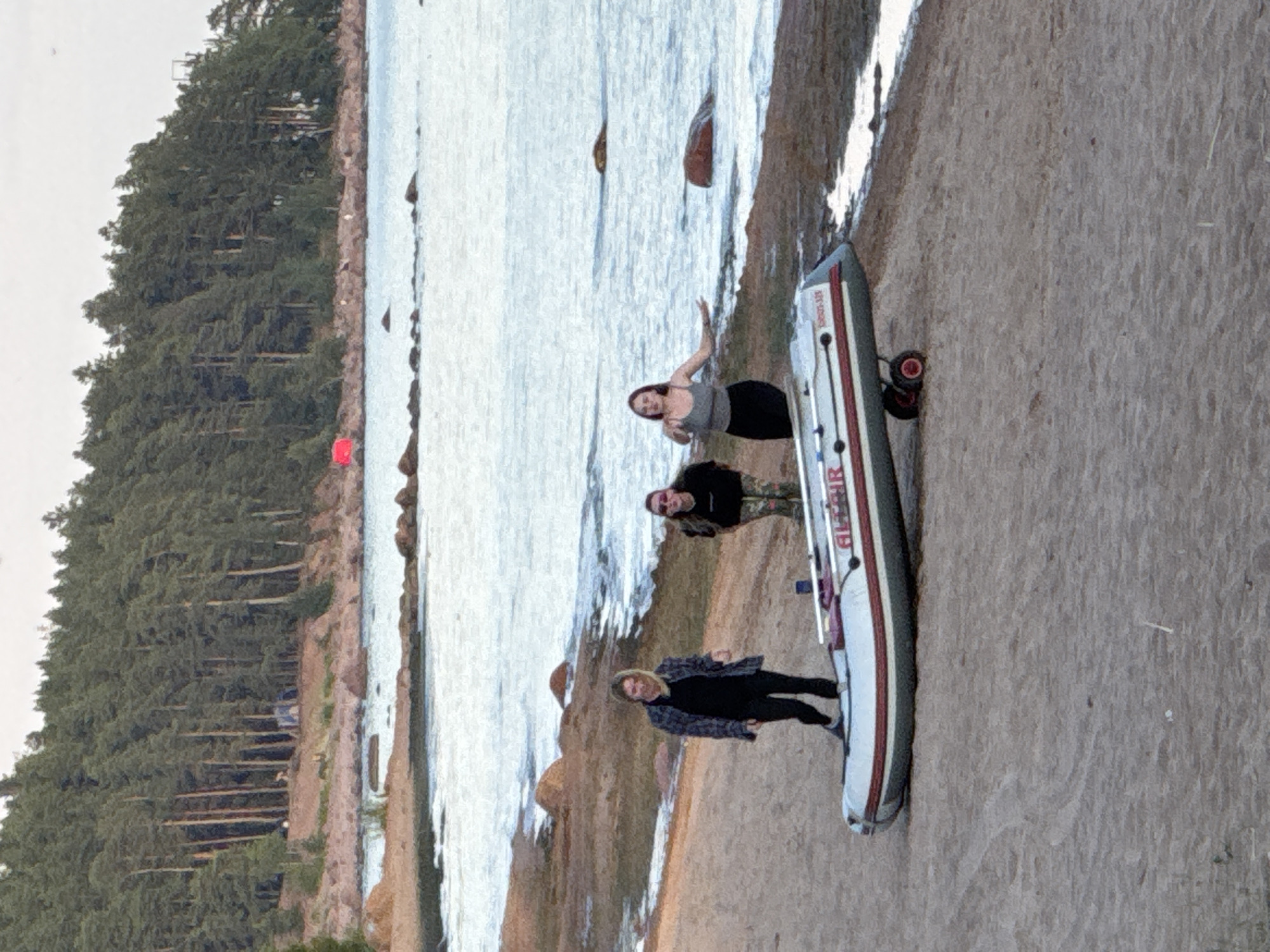 The image depicts a sandy beach area with a body of water in the background, bordered by a dense forest. Three individuals are present on the beach. In the foreground, a deflated inflatable boat with red and white accents is positioned on a two-wheeled dolly. The boat is labeled "ALRIR" and "SPORT 430".
To the left, a woman wearing a dark plaid jacket over a black shirt and black pants stands facing forward, looking towards the camera with a slight smile.
In the middle, another woman is visible, wearing a camouflage-patterned shirt and dark pants. She is looking towards the camera.
To the right, a third woman, wearing a grey tank top and a black skirt, is facing forward with her arms extended slightly outwards. She appears to be interacting with the camera.
The environment is a beach with visible footprints and some debris on the sand. The water appears calm with small waves. The forest in the background is composed of tall, dark green pine trees. Hints of tents and possibly a red flag are visible within the treeline. The sky is overcast. The image has a somewhat grainy texture, suggesting it may be a digital photograph. The location is identified as Zelenaya Roshcha, Russia, which is known for its natural coastal areas and forests.