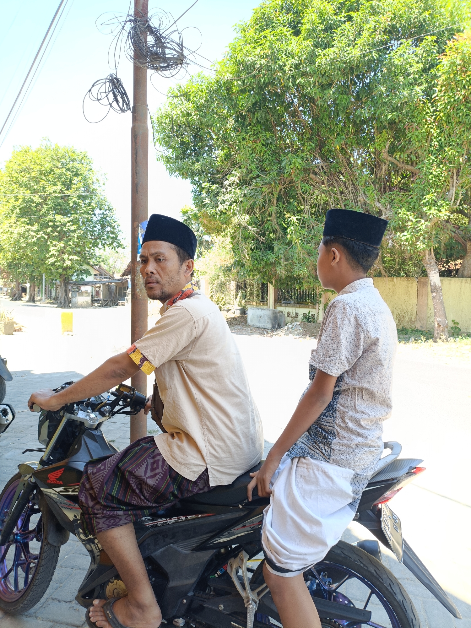 An adult male and a juvenile male, both wearing peci hats, are posed on a black motorcycle on a paved road in Pordapor, Indonesia. The adult, seated as the driver, wears a tan collared shirt with patterned trim on the collar and sleeves, and a sarong with a diamond pattern. The juvenile, seated behind the driver, wears a light-colored, patterned short-sleeved shirt and a white sarong. The motorcycle has purple accents on the wheels. In the background, a utility pole with tangled wires stands near a leafy tree. Further back, there are buildings and other trees under a clear blue sky. The lighting suggests it is daytime with bright sunlight.