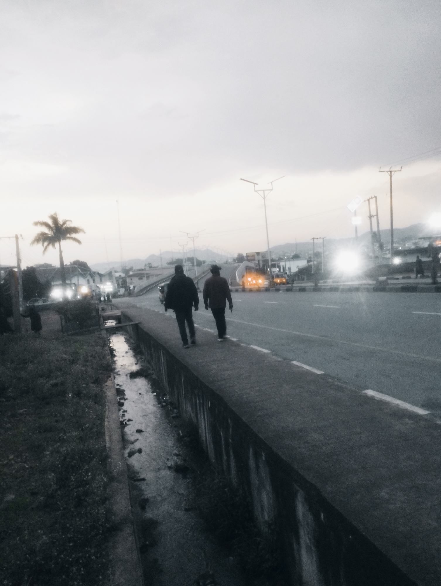 Two individuals, seen from behind, walk along a concrete sidewalk next to a narrow, shallow drainage channel filled with murky water. The sidewalk is elevated and borders a multi-lane road. In the distance, vehicles with illuminated headlights are visible on the road, along with streetlights and utility poles. A lone palm tree stands on the left side of the scene, amidst a cluster of low-rise buildings. The sky is overcast, suggesting it is either dawn or dusk. The overall impression is an urban roadside environment in Jos, Nigeria, during low light conditions.