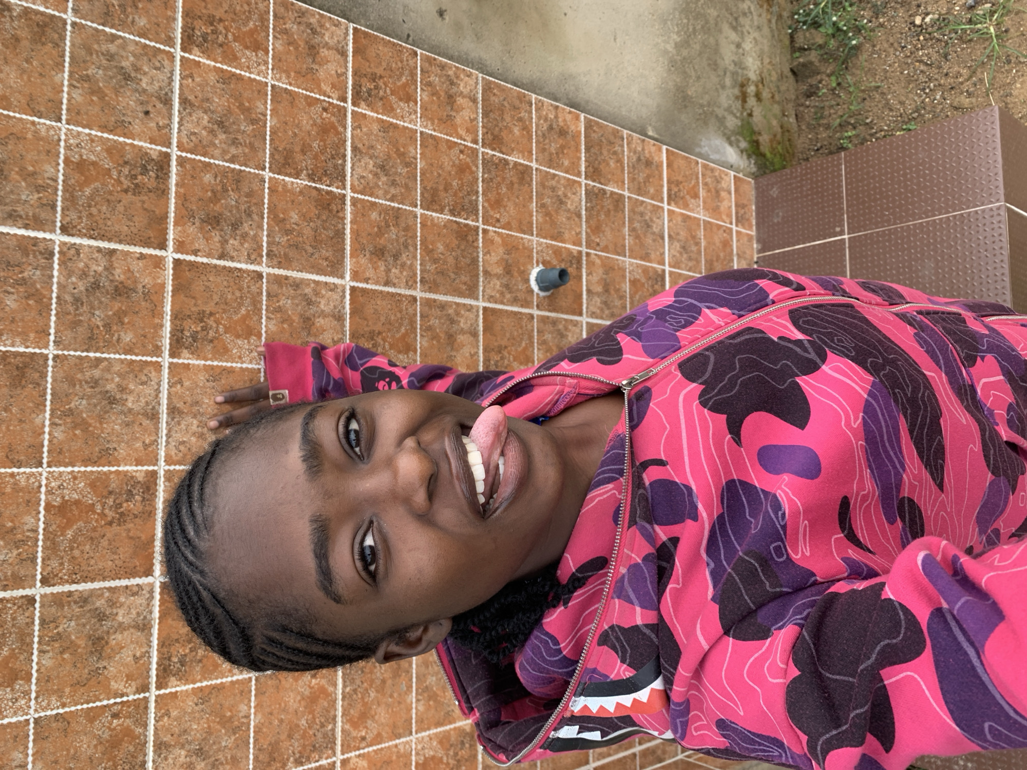 A young individual with dark skin and braided hair is captured in a waist-up, vertical photograph, presenting a playful expression with an open mouth and extended tongue, looking directly at the camera. The person is wearing a zippered hooded sweatshirt featuring a hot pink, purple, dark grey, and white camouflage pattern, accented by a prominent shark-mouth graphic on the upper left chest. Fingers of the individual's right hand are visible resting against a tiled surface in the upper right of the frame. The background consists primarily of a wall covered in mottled brown square tiles separated by decorative white, wavy grout lines. A dark grey cylindrical pipe or faucet fixture with a white end protrudes from the tiled wall. The ground transitions from a plain light grey concrete section to a darker brown tiled area with a textured surface. To the far right and bottom, an area of reddish-brown soil with sparse green vegetation is visible, indicating an outdoor or semi-outdoor location in Rafin Par, Nigeria.