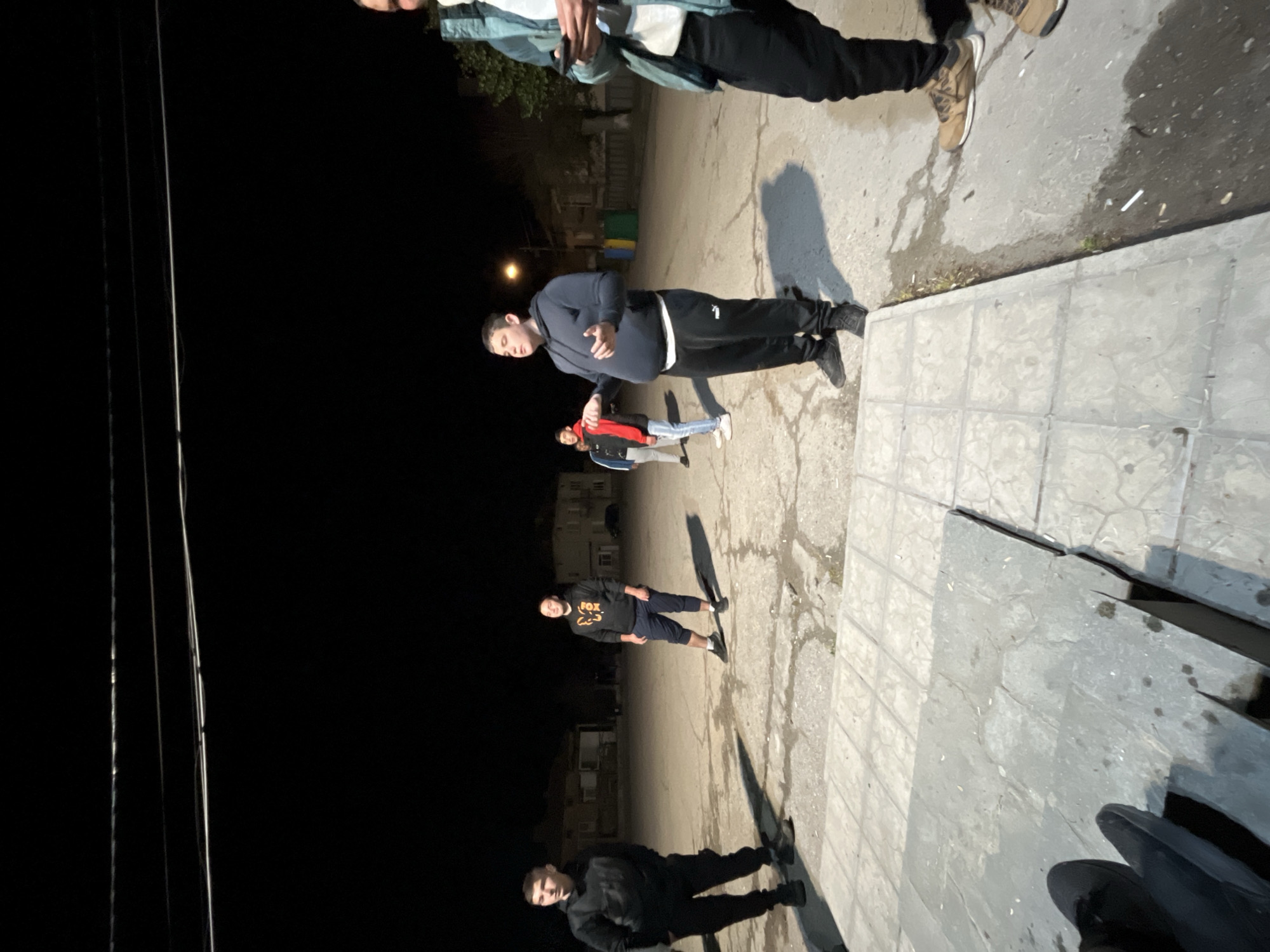 The image depicts an outdoor nighttime scene in Karavelovo, Bulgaria, showing a group of young men gathered on a paved surface. The sky is dark, indicating it is night, with artificial light sources casting distinct shadows on the ground.
In the mid-ground and center, a young man wears a dark hooded sweatshirt and black track pants with a white stripe, his hands clasped in front. To his immediate right, another person is visible from the chest down, dressed in a light teal or green jacket and dark pants. Further back, two more individuals are present; one wears a red and black jacket, while the other is partially obscured. In the background, another young man stands wearing a black hoodie with the text "FOX" visible on it, paired with dark shorts. Another figure in a dark jacket and pants is partially visible in the lower left of the frame.
The ground is a combination of rough, dark asphalt and lighter concrete paving slabs, suggesting a street, sidewalk, or public square area. Utility poles and wires extend vertically along the left side of the frame, disappearing into the darkness. Faint outlines of buildings are visible in the distant background, hinting at a residential or urban setting. The individuals appear to be standing casually, possibly socializing or waiting.