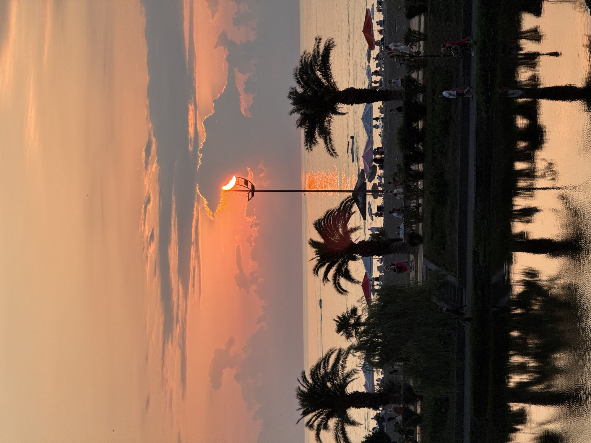 This is a photograph of a beach at sunset. The sun is setting behind a large cloud, casting a warm orange glow over the sky and water. Palm trees line the beach, and there are people sitting on lounge chairs under umbrellas. In the foreground, a body of water reflects the palm trees and the sunset. The overall impression is one of a peaceful and serene evening.