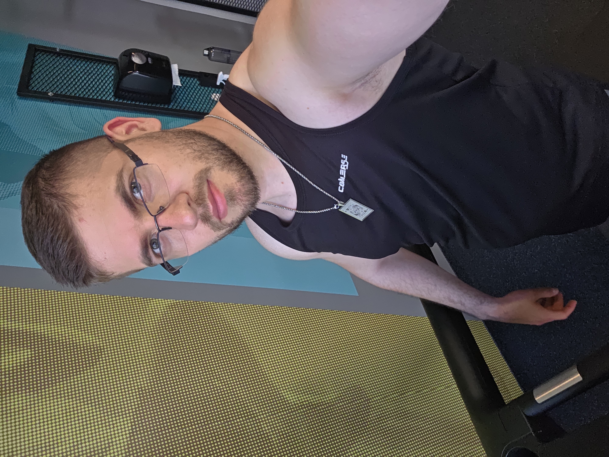 A man is taking a selfie. He is wearing a black tank top and glasses, and a silver chain with a playing card pendant. The background includes a yellow patterned wall, a blue wall, and gym equipment. There is no information to identify the location as Plovdiv, Bulgaria.