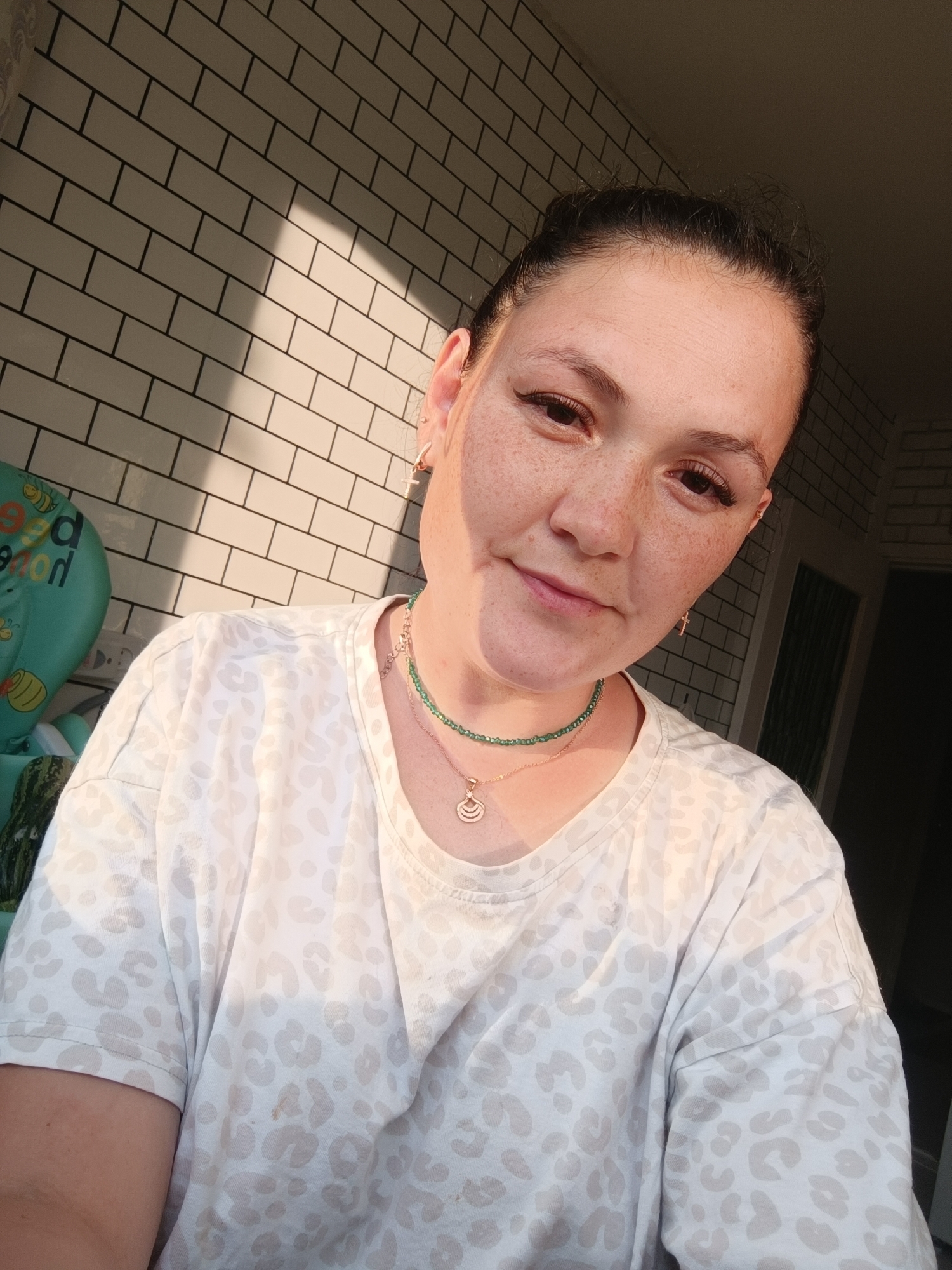 A medium shot captures a woman with dark hair pulled back, her face turned towards the camera, illuminated by warm sunlight casting a shadow on a tiled wall. She is wearing a white t-shirt with a light-colored leopard print and two necklaces. The background features a wall with white subway tiles and dark grout, and a portion of a green baby seat is visible to the left.