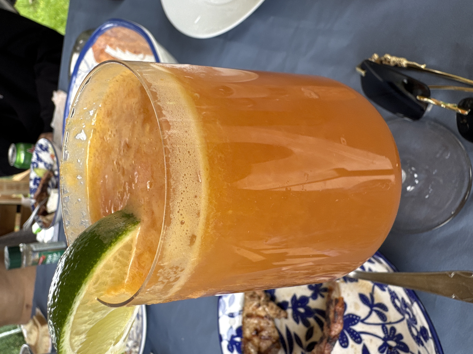 The scene is situated in Pet Kladentsi, Bulgaria, depicting an outdoor dining arrangement. In the foreground, a tall, clear stemmed glass contains a vibrant orange, frothy beverage, garnished with a fresh lime wedge positioned on the rim. The glass rests on a blue fabric tablecloth. To the bottom right of the glass, a pair of black-framed sunglasses with gold-colored temple pieces is visible. To the left of the glass, a blue and white patterned ceramic plate (with a floral/foliage design) holds several pieces of dark brown, grilled meat. A silver-colored fork is partially visible to the left of the plate. In the background, partially obscured by the main subject, a small clear shaker bottle with a green cap and a white label is on the table. To the far left, the bare shoulder and upper torso of a person with light skin are visible. To the upper right, a person wearing a black garment is partially visible, and a green aluminum beverage can is also present. A white ceramic bowl containing a lighter colored substance (possibly a dip or sauce) is visible further back on the right side. The ambient lighting suggests a daytime setting.