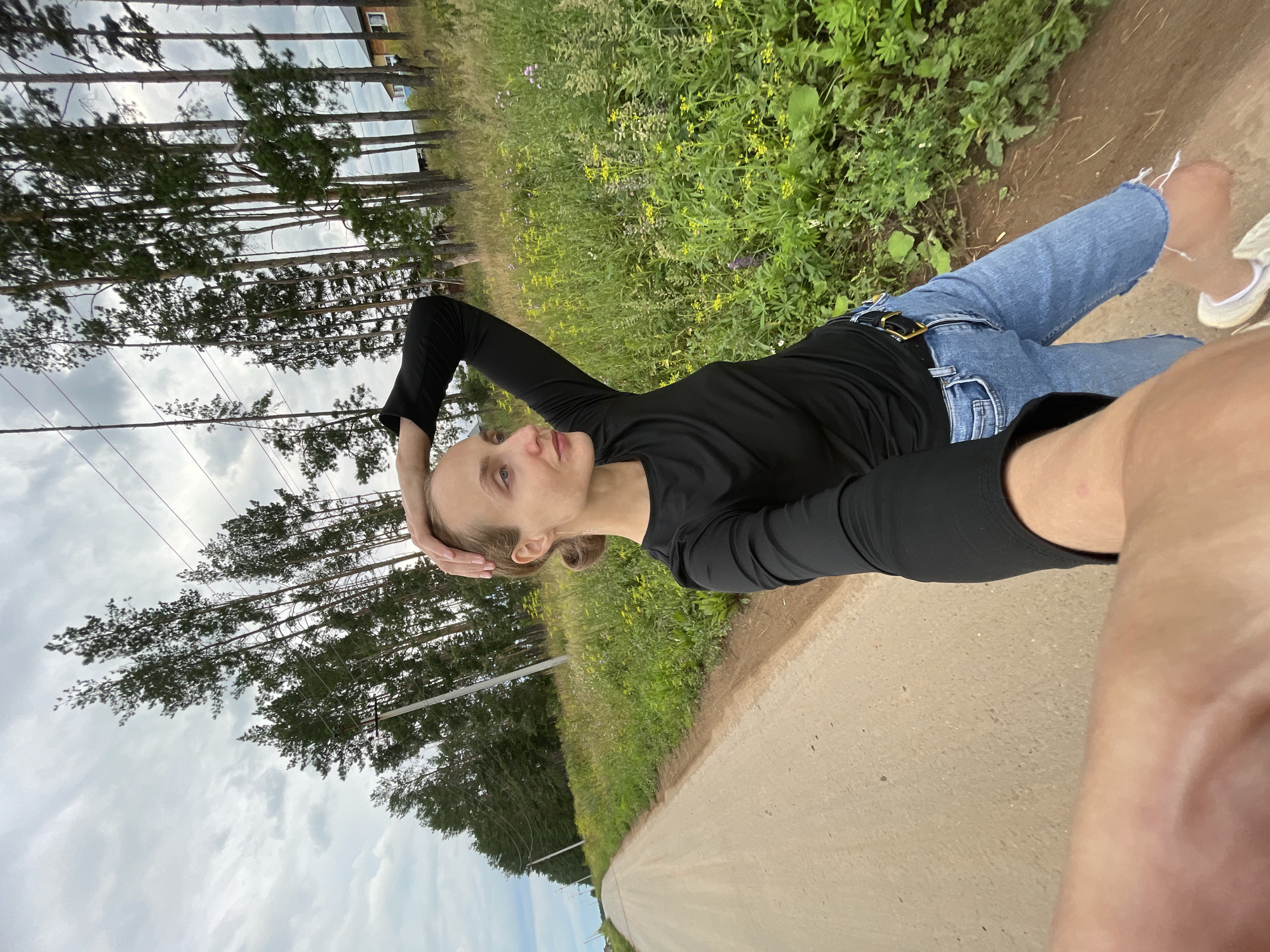 A woman with blonde hair pulled back in a bun, wearing a black long-sleeved shirt and distressed denim shorts with a black belt, stands on a dirt road in Подшивалово, Russia. She is looking to her right, with her left hand resting on the back of her head. The road stretches into the distance, lined on the right by a field of green grass and yellow wildflowers, with tall pine trees and utility poles beyond. A glimpse of a wooden house is visible through the trees in the background. The sky is overcast with clouds. The photograph is taken from a low angle, with the woman's arm and hand in the foreground.