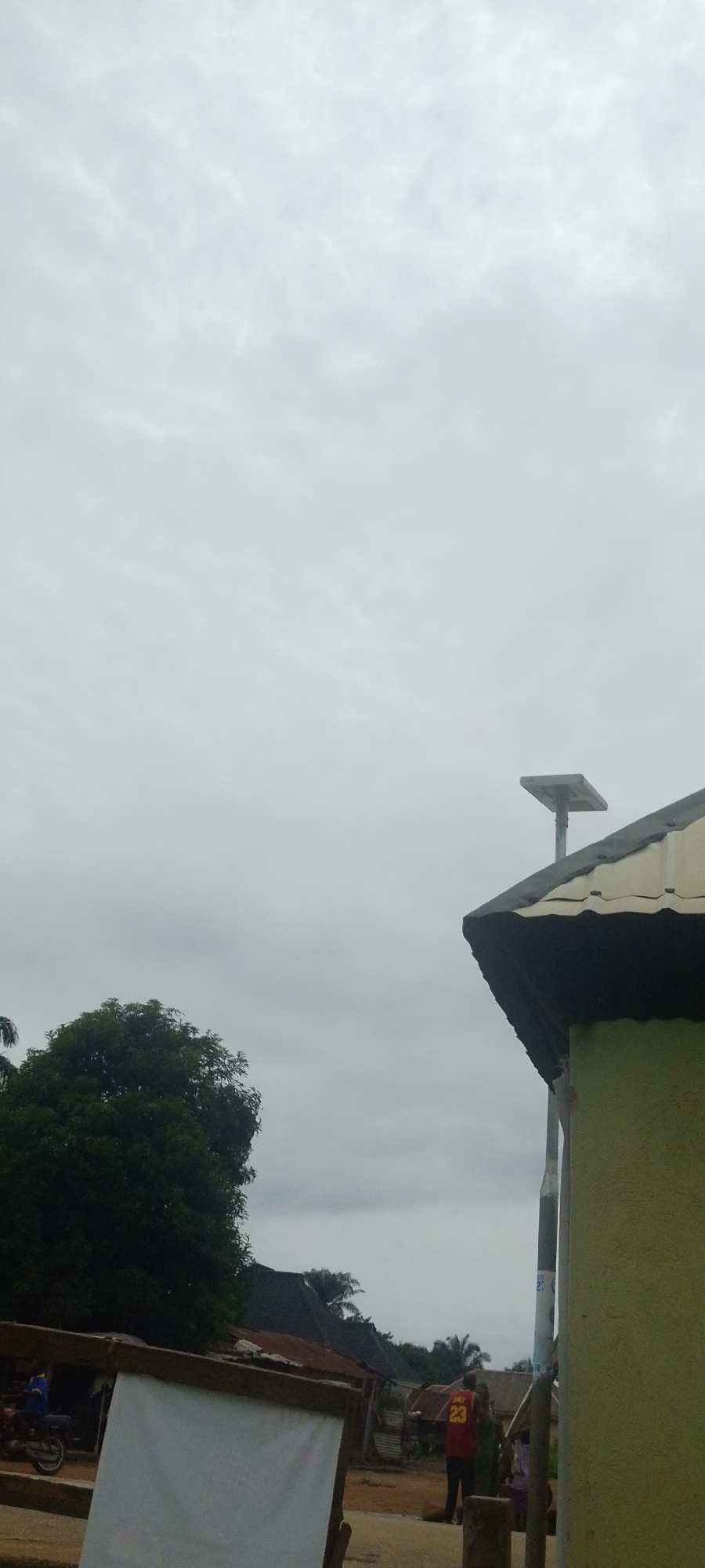 The image depicts an overcast sky over a semi-urban or rural setting in Baissa, Nigeria. In the foreground, a white, rectangular object, possibly a banner or sign, is partially visible, supported by wooden beams. To the left, a dark green, dense tree is prominent. Behind the tree, several buildings with corrugated metal roofs are visible. To the right, the corner of a building with a yellow-green wall and a dark roof overhang is in focus. A pole with a square solar panel mounted on top extends upwards from the roof. Further into the scene, a person wearing a red jersey with the number "23" is standing, facing away from the camera. Other individuals are also present in the background, near the buildings and what appears to be a dirt road. A motorcycle is parked to the left of the white banner. The overall atmosphere suggests a typical day in this Nigerian locality.