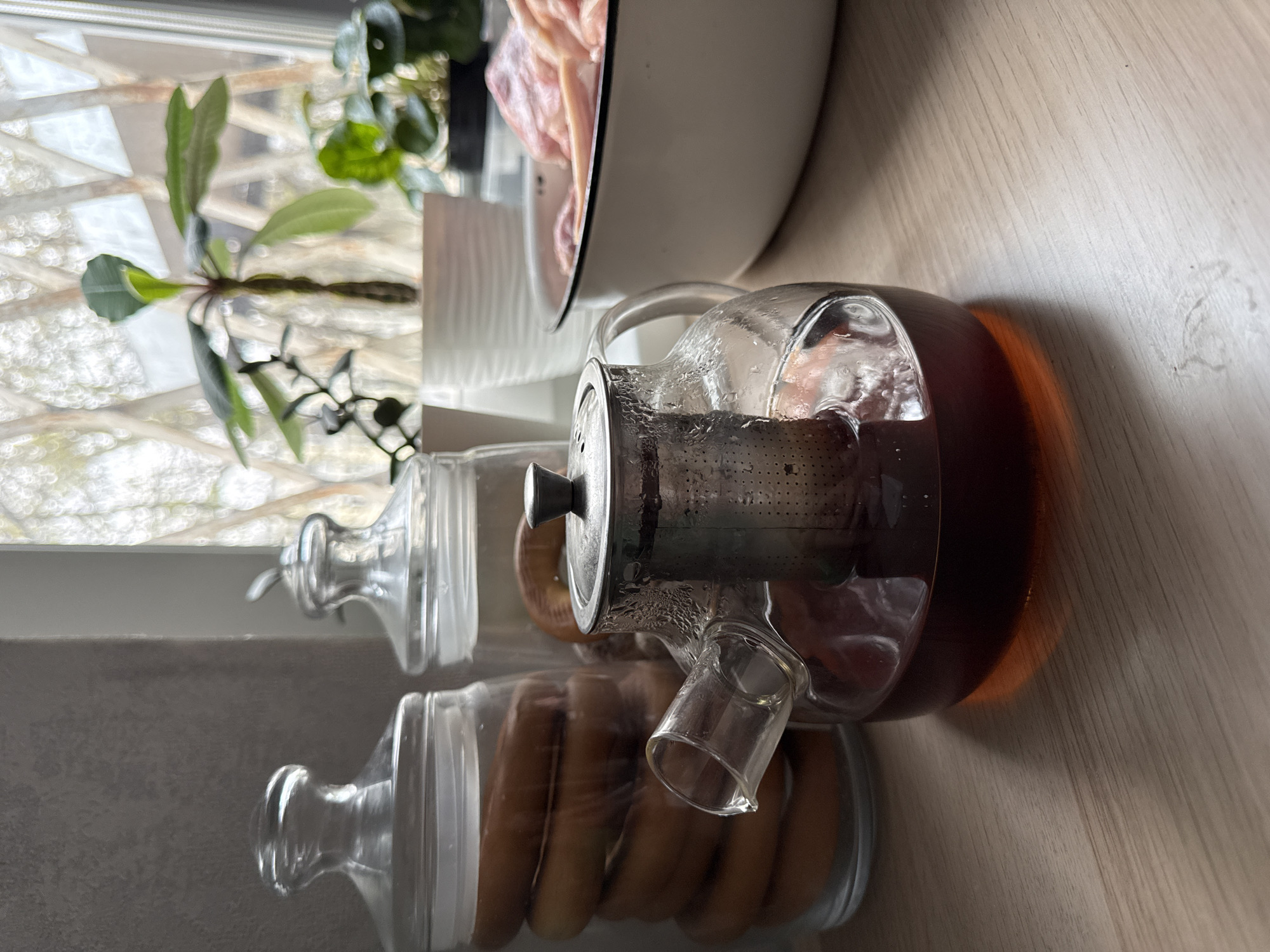 A clear teapot filled with a dark liquid, likely tea, sits on a light brown wooden surface. To the left of the teapot, two clear glass jars with lids contain cylindrical items that appear to be bagels or breadsticks. Behind the jars is a window with a blurred view of trees and foliage, suggesting an outdoor environment. A small plant is also visible near the window. In the background, a white bowl, partially filled with meat, sits next to the window. The scene appears to be indoors, possibly a kitchen or dining area, but there is no confirmation of it being in Moscow, Russia.