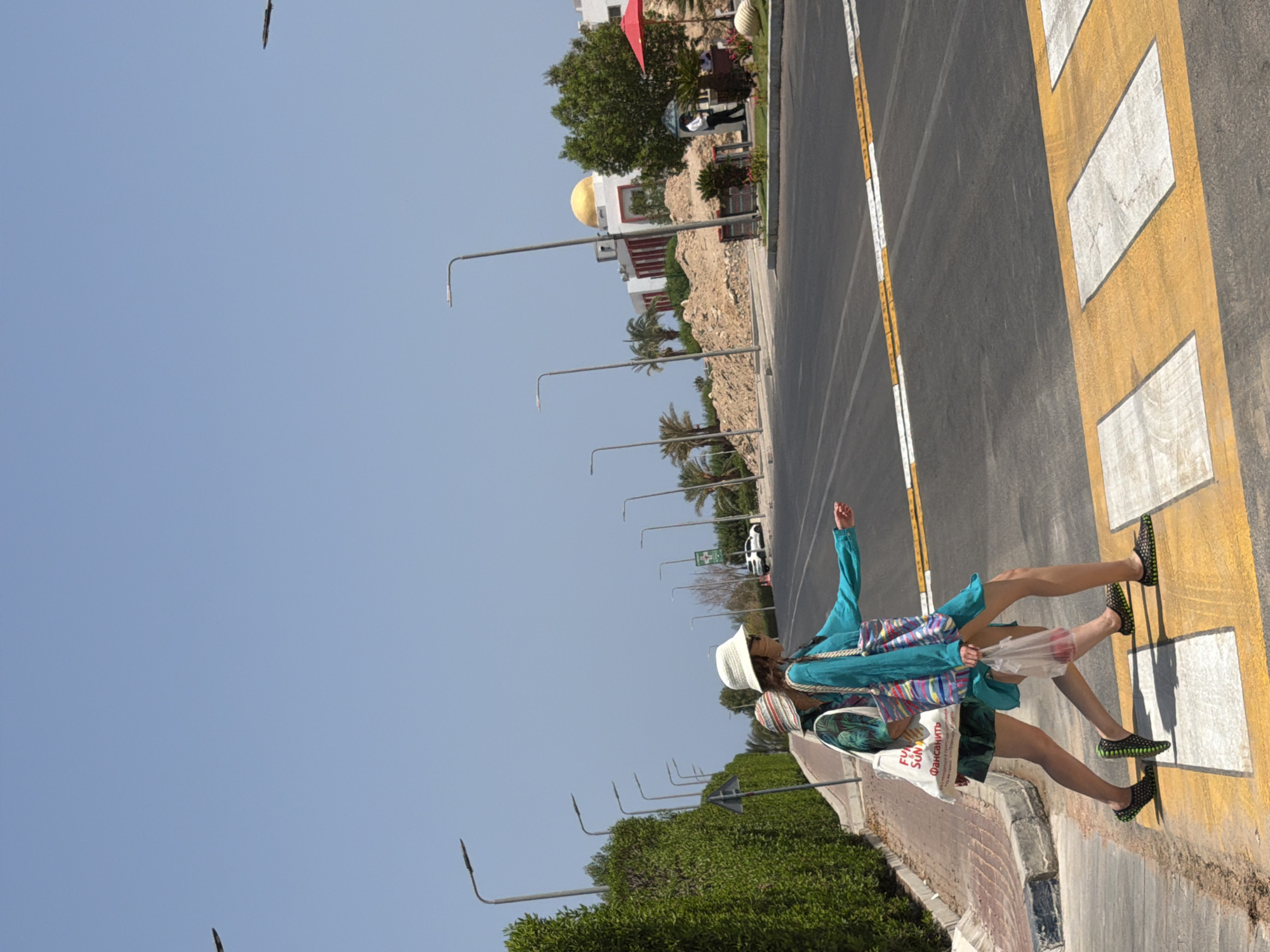 Two females, one wearing a teal dress and white sunhat, the other in a green patterned shirt and multi-colored bucket hat, cross a street at a pedestrian crossing. They are walking from left to right across the frame. The woman in the teal dress carries a clear plastic bag with red items inside. Both are wearing dark mesh shoes with bright green accents. The woman in the teal dress is reaching out her right hand.
The pedestrian crossing has yellow and white stripes. The street is asphalt. To the left of the street, there is a sidewalk and a dense green hedge. Beyond the hedge, streetlights are visible. In the background, across the street, are buildings with white facades and a gold dome, palm trees, and construction materials (piles of dirt and rubble). A red umbrella is visible further in the background. The sky is clear and blue. The location is identified as Sharm El Sheikh, Egypt, with elements such as palm trees, arid construction areas, and the architectural style of the buildings being indicative of the region.