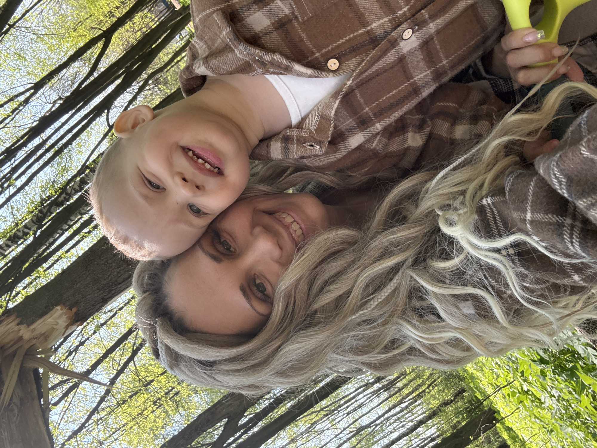 The image shows a woman and a young child outdoors. They appear to be in a wooded area, possibly a park, evidenced by the presence of trees and green foliage. The child has blonde hair and is wearing a brown plaid shirt. The woman has long blonde hair and is also wearing a brown plaid shirt. Both are smiling and appear to be enjoying themselves. The location is specified as Rusanovo, Russia, although there are no specific identifying landmarks to confirm this location.