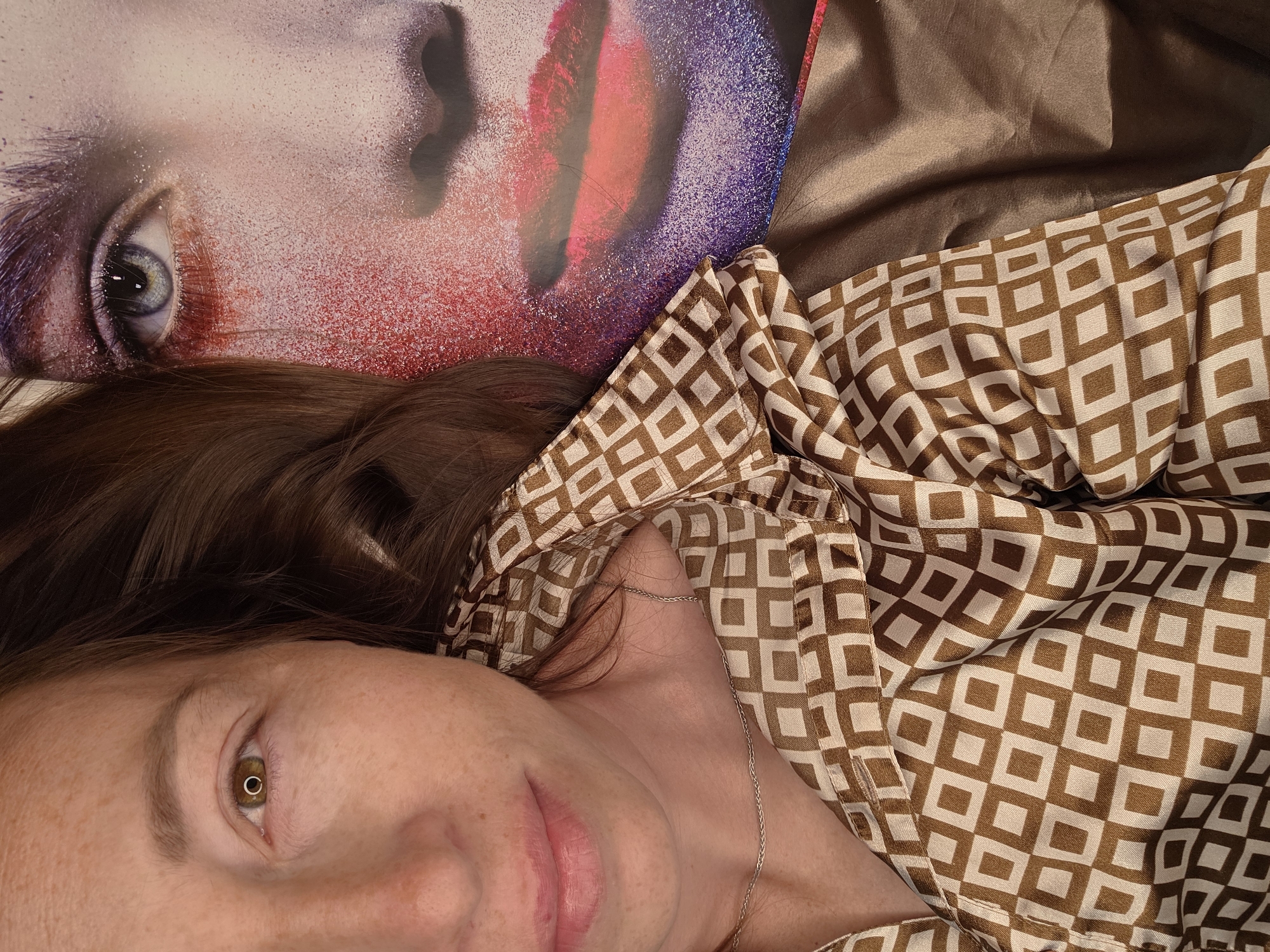 The image shows a close-up, overhead view of a person lying down with their head turned towards the camera. They are wearing a brown and white patterned button-down shirt and a silver necklace. To the right of the person's face is a large, colorful image of a person's face with dramatic makeup, featuring bright red and purple eyeshadow and bold red lipstick. The background appears to be a flat surface, possibly a bed or a floor, with a sheen to it, suggesting it could be a silk or satin material. The lighting is soft, creating a gentle illumination on the person's face. The scene is indoors, and there are no visible windows or outdoor elements.