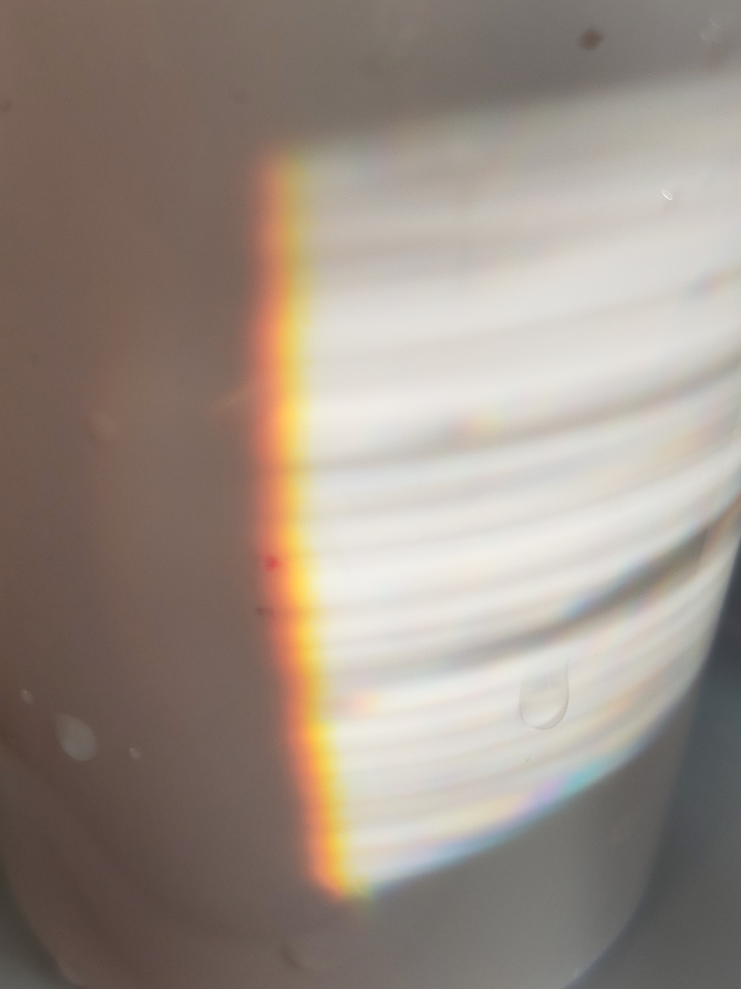 The image is a close-up, blurry view of a white, ridged cylindrical object, possibly a cup or vase, with a rainbow prism effect on the surface. There are water droplets or imperfections visible on the object's surface. The image does not provide sufficient information to place it in a specific location, city, or country. There are no people present.