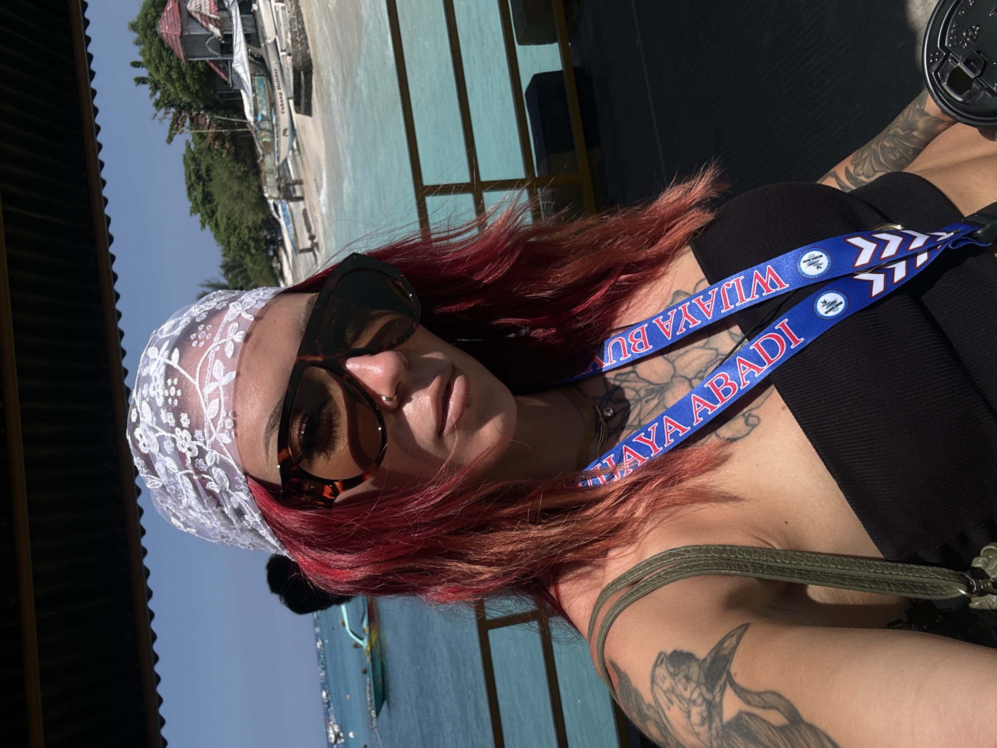 A medium shot shows a person with red hair wearing a white lace head covering and tortoise-shell sunglasses. The person also wears a blue lanyard with the words "WIJAYA ABADI" printed on it. They have visible tattoos on their chest and arm. The background depicts a beach with boats and buildings, indicating a coastal area. The location is unknown, but the lanyard suggests it is in Indonesia. The photo is taken outdoors during daylight.