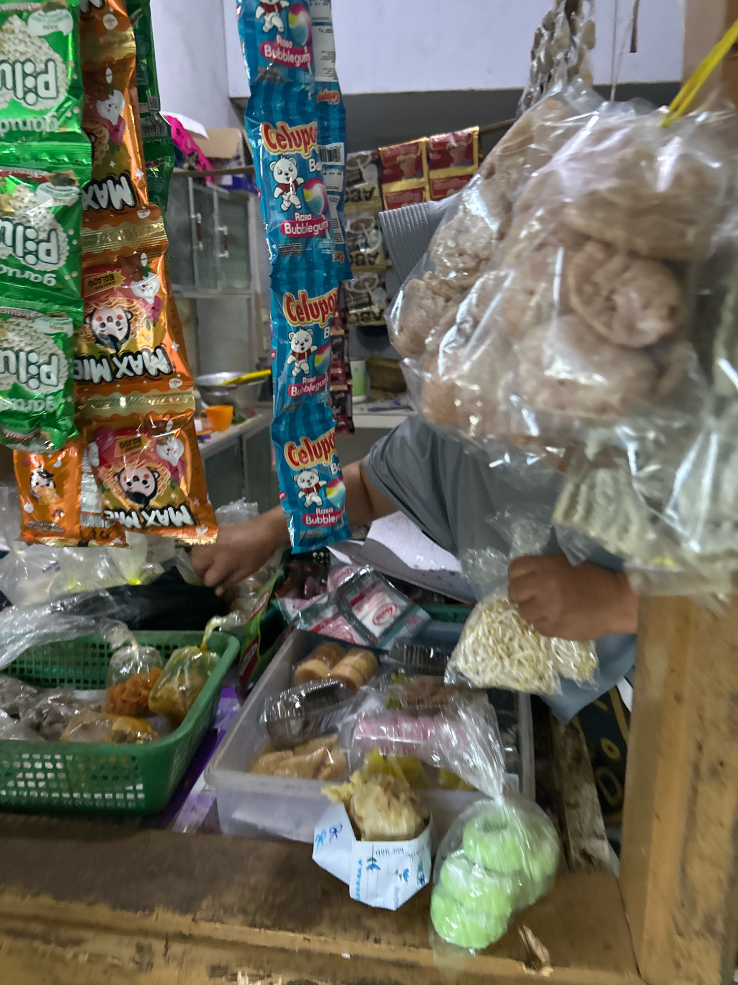 The image depicts a close-up view of a small market stall, likely in Cihaurbeuti, Indonesia. The foreground is dominated by a wooden counter with various food items. In the center, a green plastic basket overflows with small packaged snacks and fried items. To the right of the basket is a clear plastic container holding more food, including what appear to be small pastries or cakes. In the background, a person wearing a grey t-shirt is visible, their hands engaged in arranging or handing over a bag of white noodles or sprouts. Hanging from above are several rows of brightly colored snack packets, including green and orange packages with prominent text like "MAX MIE" and blue packets with images of a bear and text indicating "Celupos" and "Rasa Bubblegum." A stack of large, light-colored, crispy snacks is also visible, packaged in clear plastic. The overall environment suggests a bustling, local commerce setting with a variety of packaged and bulk food products on display.