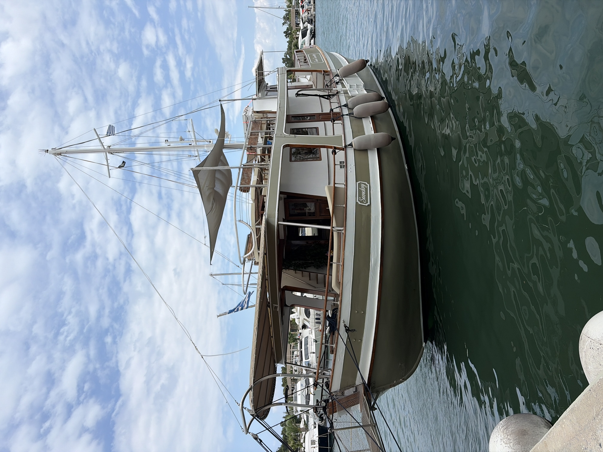 A motor yacht named "Sunday" is moored in a harbor in Kassandra Municipal Unit, Greece. The yacht is white and green with tan awnings and a wooden deck. It has a tall mast with a Greek flag. Several fenders are hanging from its side. The water is calm with reflections of the boat and the sky. Other boats are visible in the background, as well as trees and buildings along the shoreline. The sky is blue with scattered white clouds. The image is taken from a low angle, showing the boat from the waterline up.
