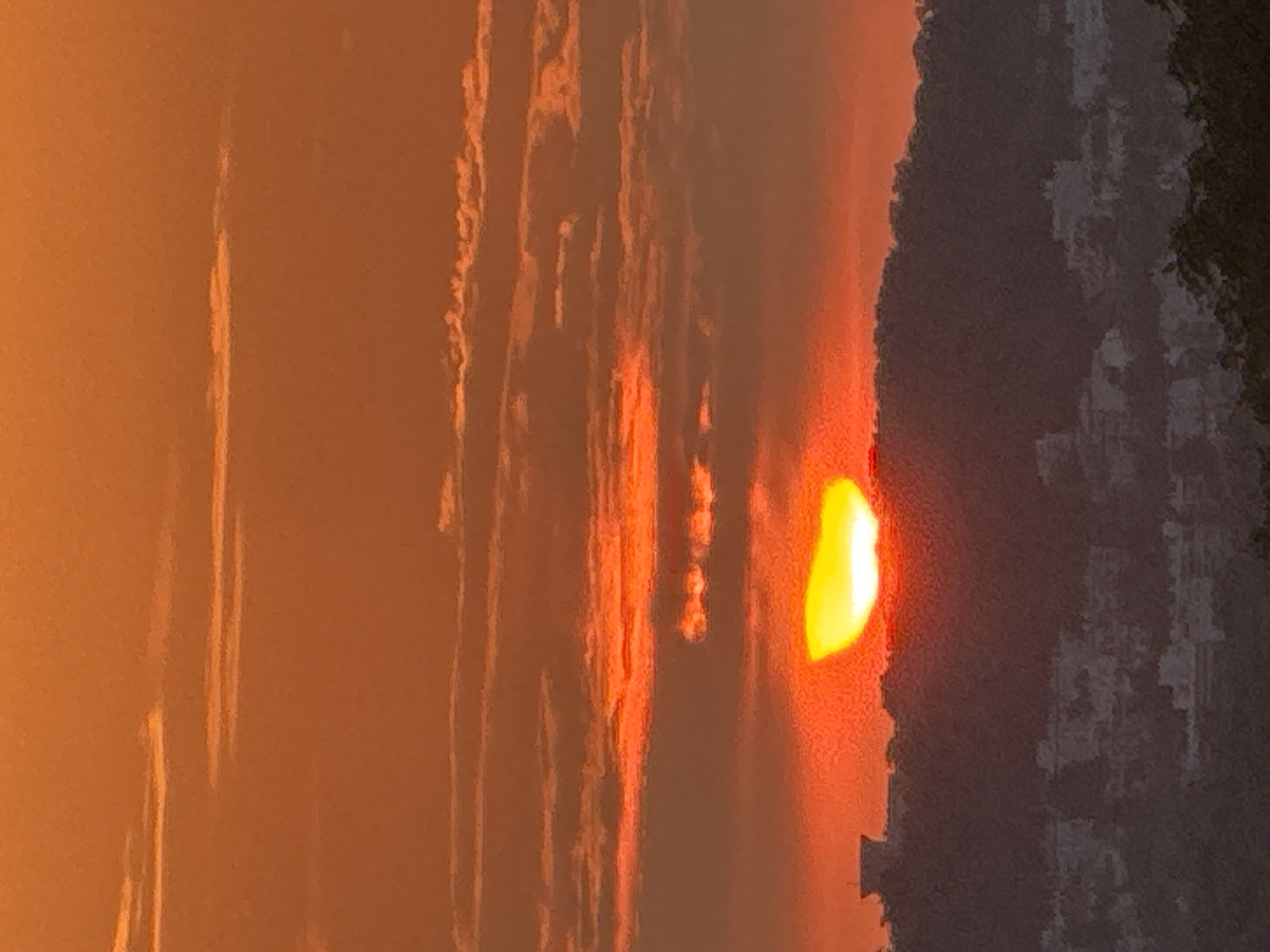 The image captures a vibrant sunset over a silhouetted landscape. The sky is a brilliant orange, with streaks of darker clouds intermingling with lighter, illuminated ones. The sun, a bright yellow-orange crescent, is just above the horizon, casting a warm glow. A dark, hilly silhouette stretches across the middle ground, with the faint outlines of buildings visible on its slopes and a prominent, block-like structure on the left peak. In the foreground, the lower portions of trees are visible against the dusky atmosphere. The overall impression is one of a serene and colorful end to the day.