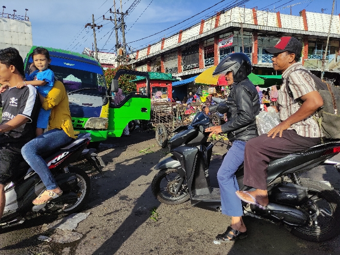 The scene is a street in Kecamatan Lawang, Indonesia (latitude: -7.83, longitude: 112.697). There are three people, two men and one young girl, and two motorcycles, one parked and one being ridden. The girl is wearing blue clothing and is sitting on the lap of the man on the parked motorcycle. The man on the parked motorcycle is wearing a black t-shirt and jeans, holding the girl. He is sitting on a black motorcycle with a red and white stripe. The man on the motorcycle that is being driven is wearing a black leather jacket, blue jeans, and a helmet. He is driving a black motorcycle. The second man is wearing a striped shirt and khaki pants. He is sitting on the back of the motorcycle being driven. There is a green truck parked behind the parked motorcycle. The street is paved. There are buildings and other vehicles in the background. The day is clear.