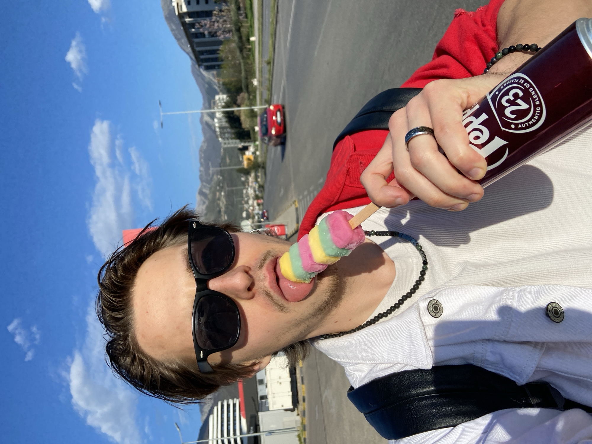 A young adult male is holding a multicolored popsicle with his tongue extended toward the popsicle and a dark-colored can that is being held in his other hand. The can is labeled "Pepper 23 Blend of 23 Flavors Authentic". The man is wearing black sunglasses, a black beaded necklace, a white denim jacket over a white t-shirt, and a red hooded sweatshirt. He has a silver ring on his left hand and a beaded bracelet on his right wrist. A black strap that appears to be a backpack is visible over his left shoulder. The background shows a paved area, a roadway with a red car, buildings, and mountains in the distance. The sky is blue with scattered clouds. The location is Bar, Montenegro.