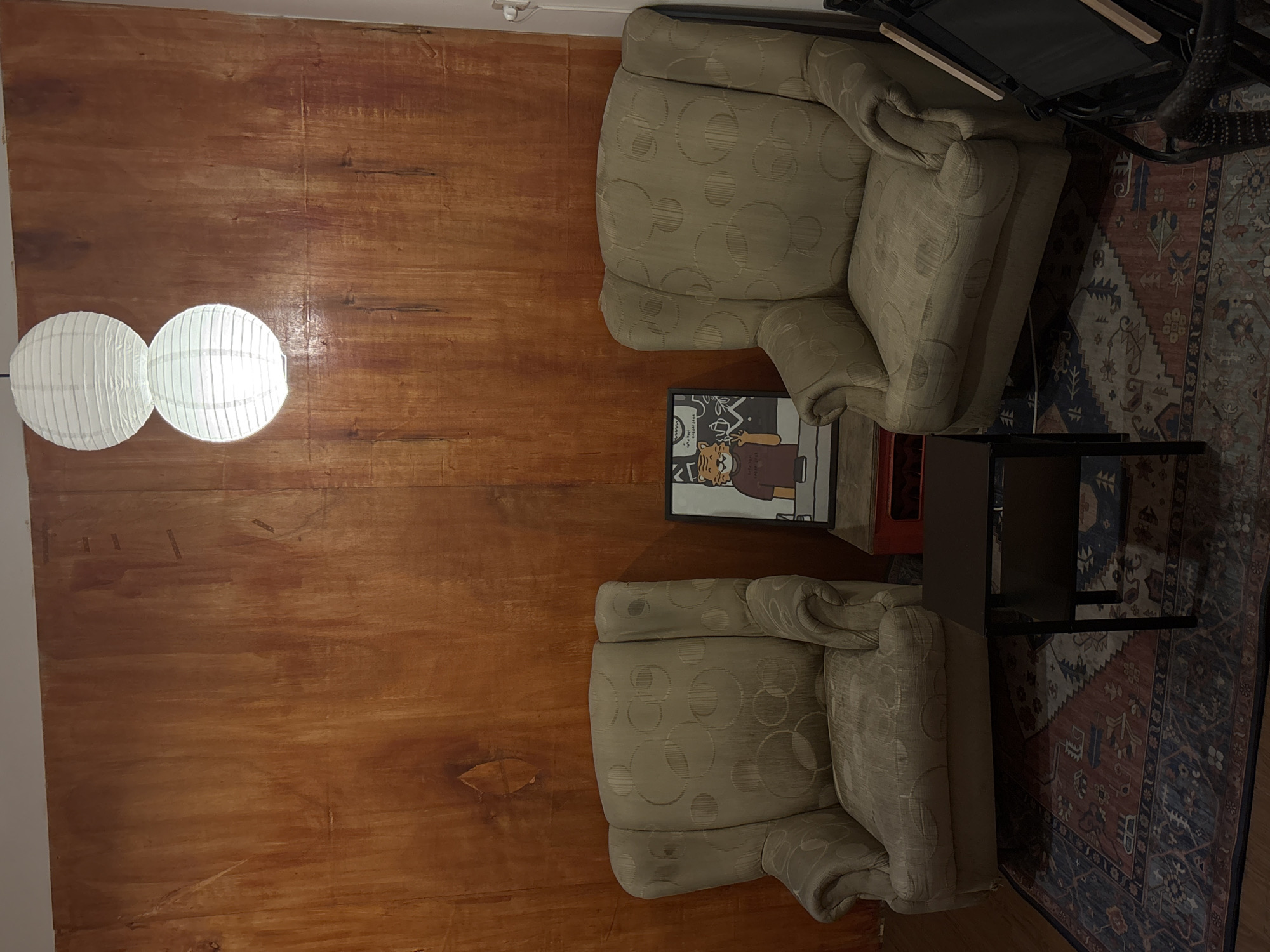 The image depicts a dimly lit interior scene featuring two armchairs, a small coffee table, and a patterned rug. The armchairs are upholstered in a beige fabric with a subtle circular pattern. A framed picture of a cartoon tiger is placed on top of a red crate, between the two chairs. Above the chairs, two white paper lanterns are suspended from the ceiling, emitting a soft glow. The background is a large, wooden panel with a reddish-brown hue, showing variations in grain and color. The overall impression is one of a cozy, possibly vintage, seating area. There are no individuals present in the image.
