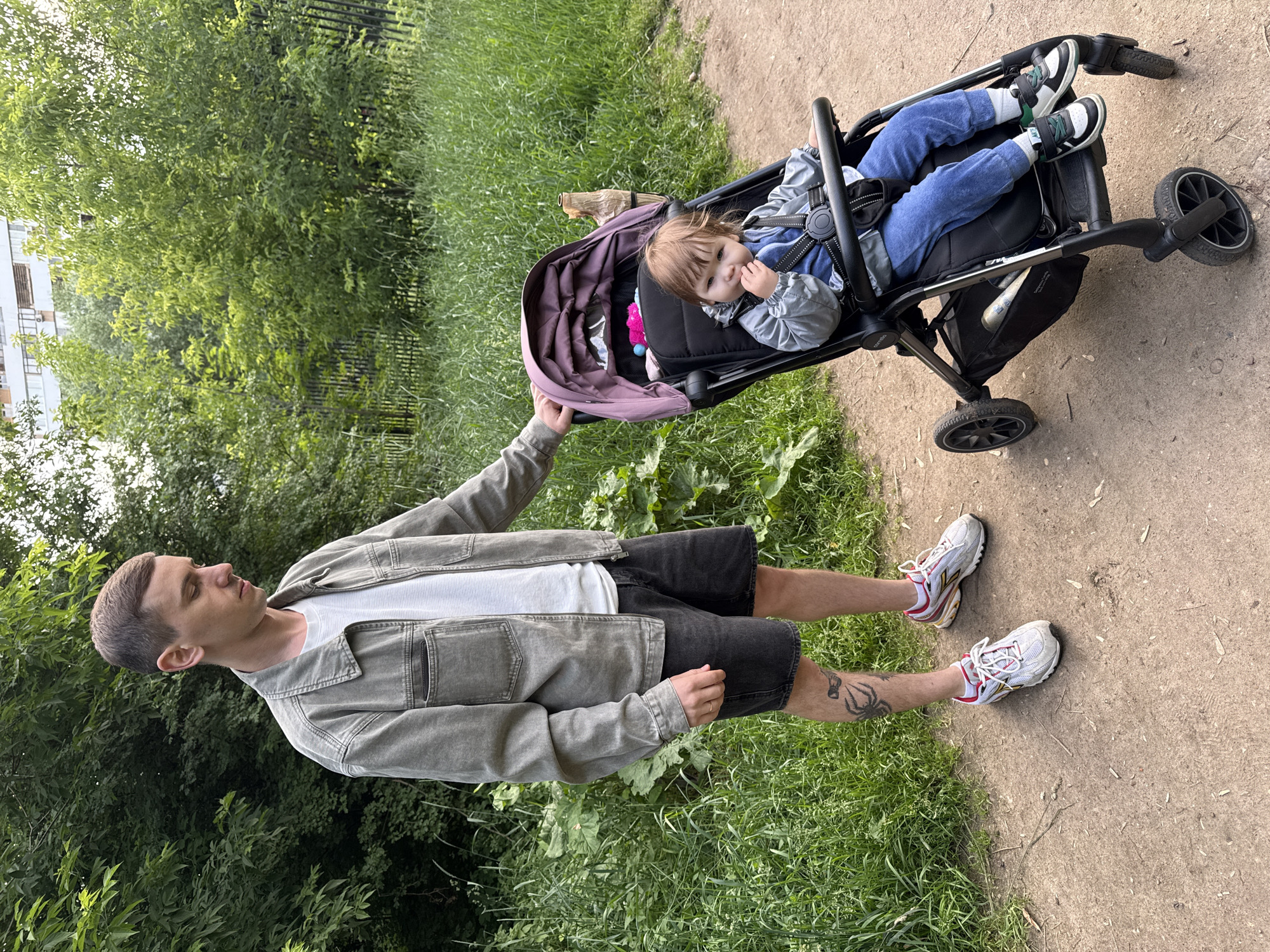 A young man stands on a dirt path in a park, his left hand resting on the handle of a stroller. The stroller contains a toddler wearing a gray jacket and blue pants, who is looking down and appears to be eating something. The man is wearing a gray jacket, a white t-shirt, and black shorts. He has a tattoo on his left leg and is wearing white and red athletic shoes. The background consists of lush green trees and grass, with a building visible in the distance. This scene is consistent with a park environment in Moscow, Russia, likely during the warmer months.