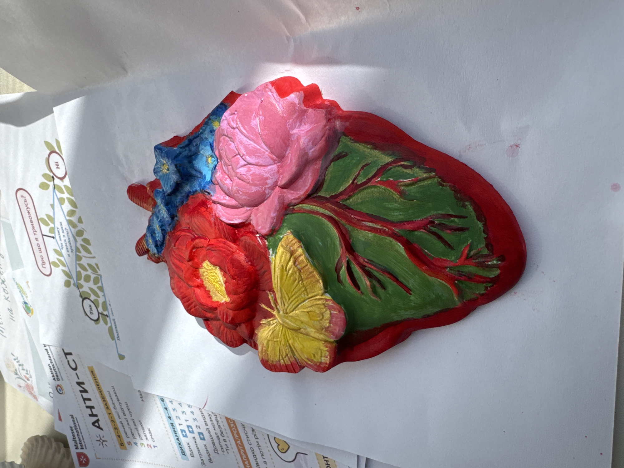 The image shows a heart-shaped sculpture decorated with flowers and a butterfly, placed on top of several sheets of paper. The sculpture is painted with bright colors: red for the heart outline, pink for a large rose, red for another flower with a yellow center, blue for smaller flowers, green for a leaf, and yellow for a butterfly. The papers beneath the sculpture appear to have text and diagrams in a Cyrillic alphabet. The paper on top has the words "АНТИ-СТ" visible. The location of Kryvyi Rih, Ukraine is not discernible from the image.