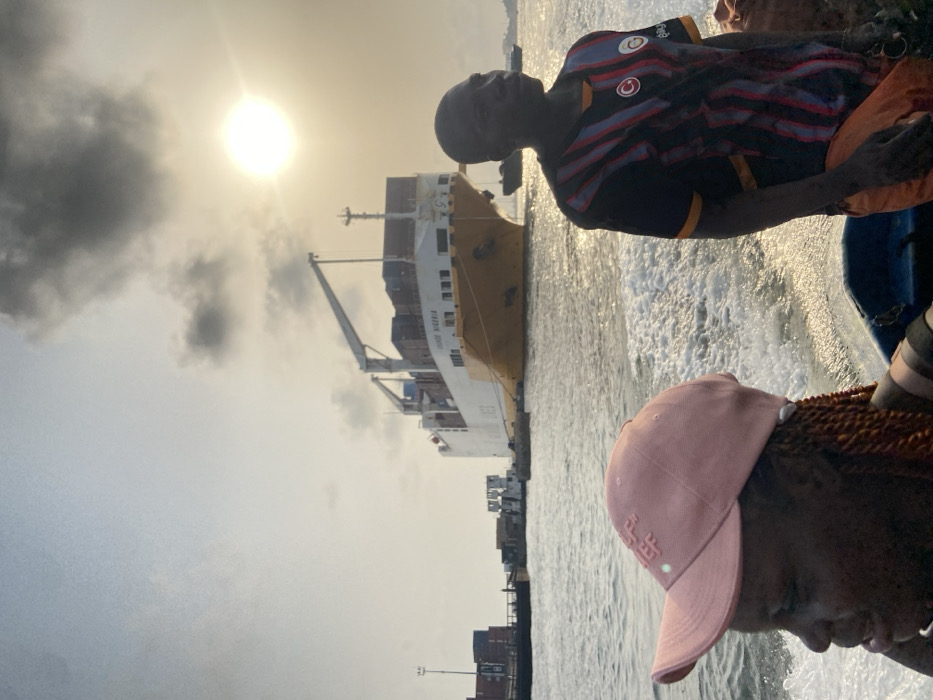 A cargo ship named “Triada Vistoria” is visible on the water near a port. People are on a boat, looking towards the vessel. The sky is overcast, with a bright sun shining through. One person wears a pink hat, while another has a black, red, and white striped shirt. The water is choppy, indicating movement. Shipping containers are stacked in the port area.