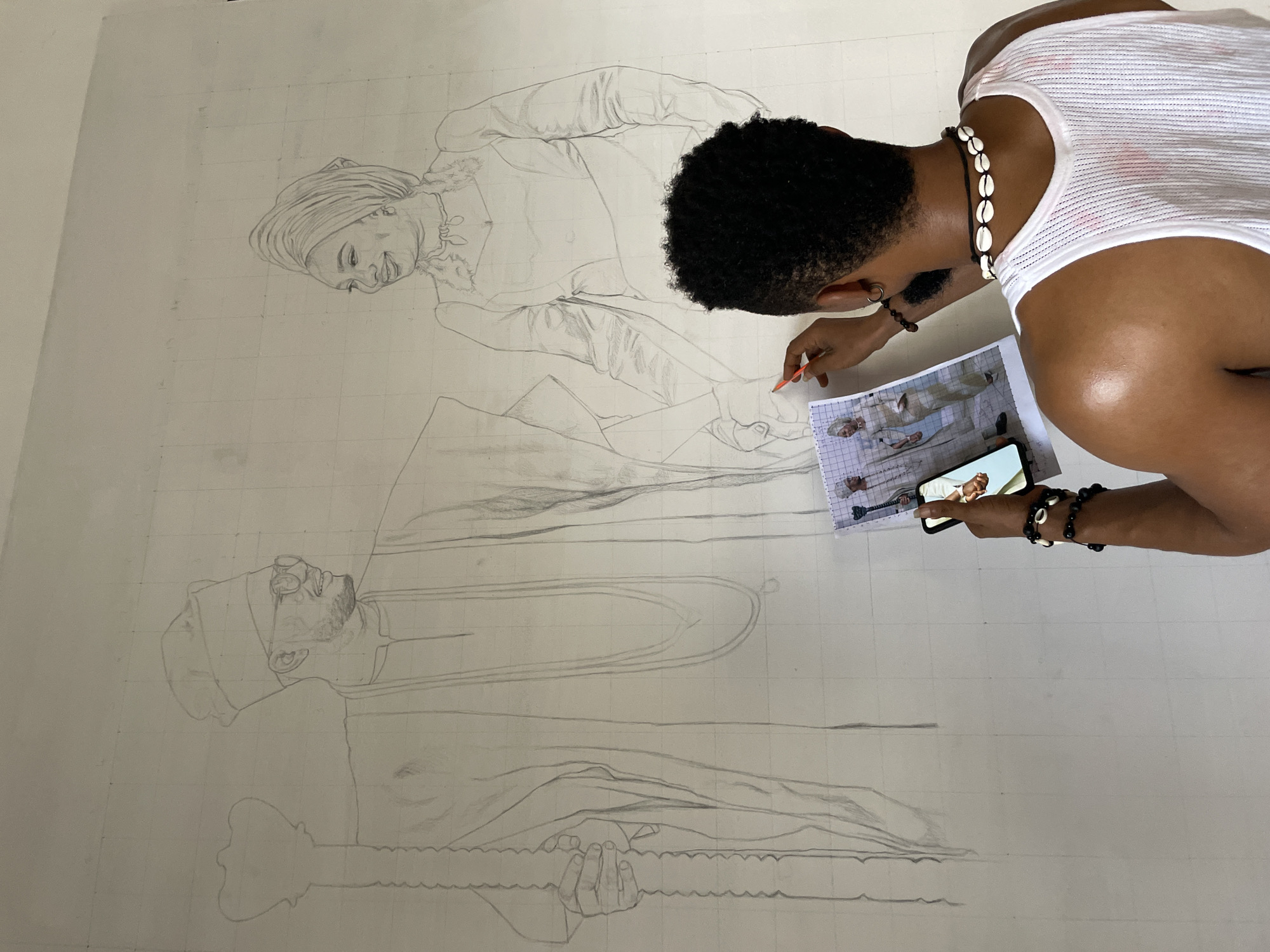 A man wearing a white tank top, shell necklace, and bracelets is drawing on a large wall in Ado, Nigeria. The wall is covered with a grid, and the sketch depicts two figures: an older man with a hat holding a wavy object and a younger woman looking down. The man is using a pencil and referencing a photo on his phone and a small printed version of the same photo. The room appears to be indoors and brightly lit.