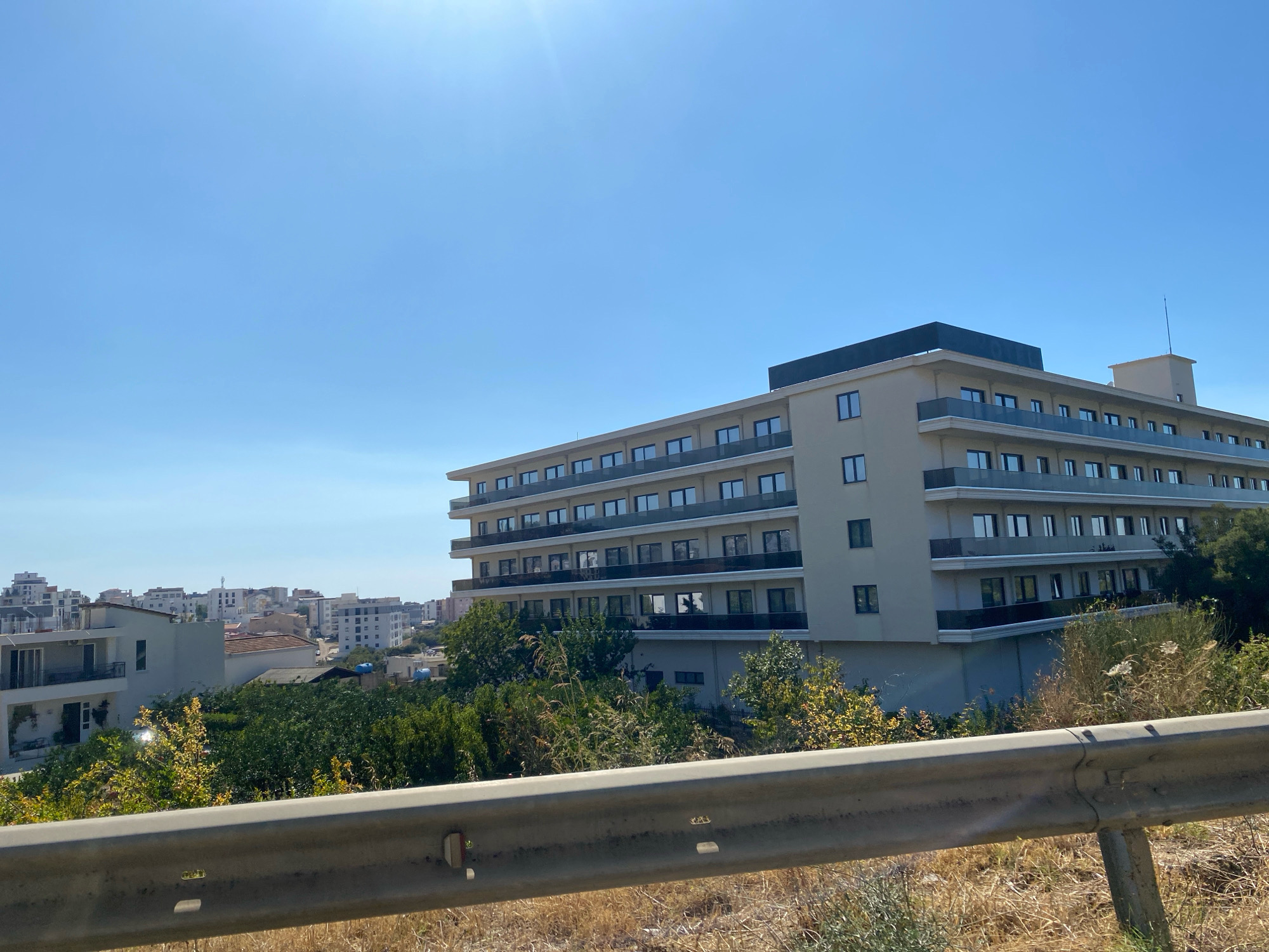 The image shows a modern, multi-story building with a light-colored facade and multiple balconies with dark railings. The building is situated on a slight incline, with a clear blue sky and some scattered clouds visible above. In the foreground, there is a silver metal guardrail with dry grass and vegetation underneath it. Behind the main building, there are other smaller buildings and some greenery, suggesting a suburban or semi-urban setting. The overall scene appears to be a sunny day. The location is identified as Dobra Voda, Montenegro.