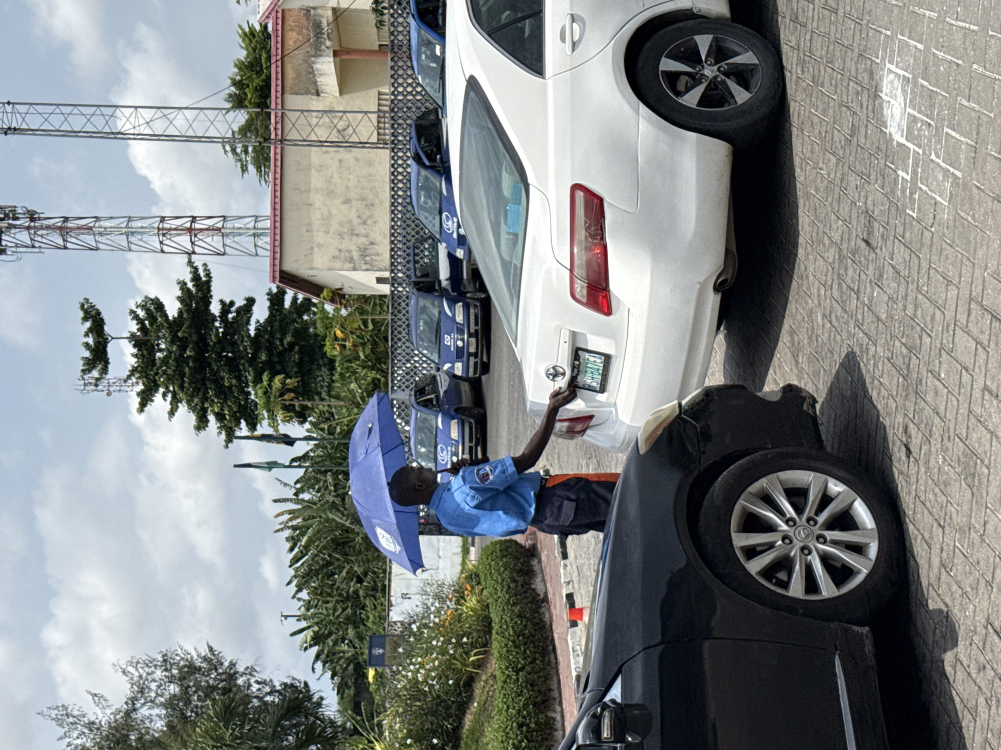This image, taken in Lekki, Nigeria, captures an outdoor scene on a bright, sunny day with a partly cloudy sky.
The primary subject is a man standing between two cars, holding a blue umbrella open over his head to shield himself from the sun. He is wearing a light blue uniform shirt with dark trousers and orange reflective stripes on the sides of his legs. A logo patch is visible on his right sleeve. He is facing a white sedan and appears to be interacting with its rear license plate, possibly cleaning it or performing a check. The license plate clearly displays "SH 847EJ".
To the right in the foreground, part of a black car is visible, including its wheel. Behind the white sedan, a row of identical blue cars is parked. These vehicles appear to be service cars, possibly taxis or company vehicles, as the word "SKYTEL" is discernible on the side of one of them.
The ground is paved with interlocking blocks, and white lines mark parking spaces. In the background, there is abundant green foliage from trees and bushes, including a well-maintained hedge. A light-colored building with a reddish roof is partially visible, and two tall metal communication towers rise into the sky. The distinct shadows cast on the ground suggest it is around midday.