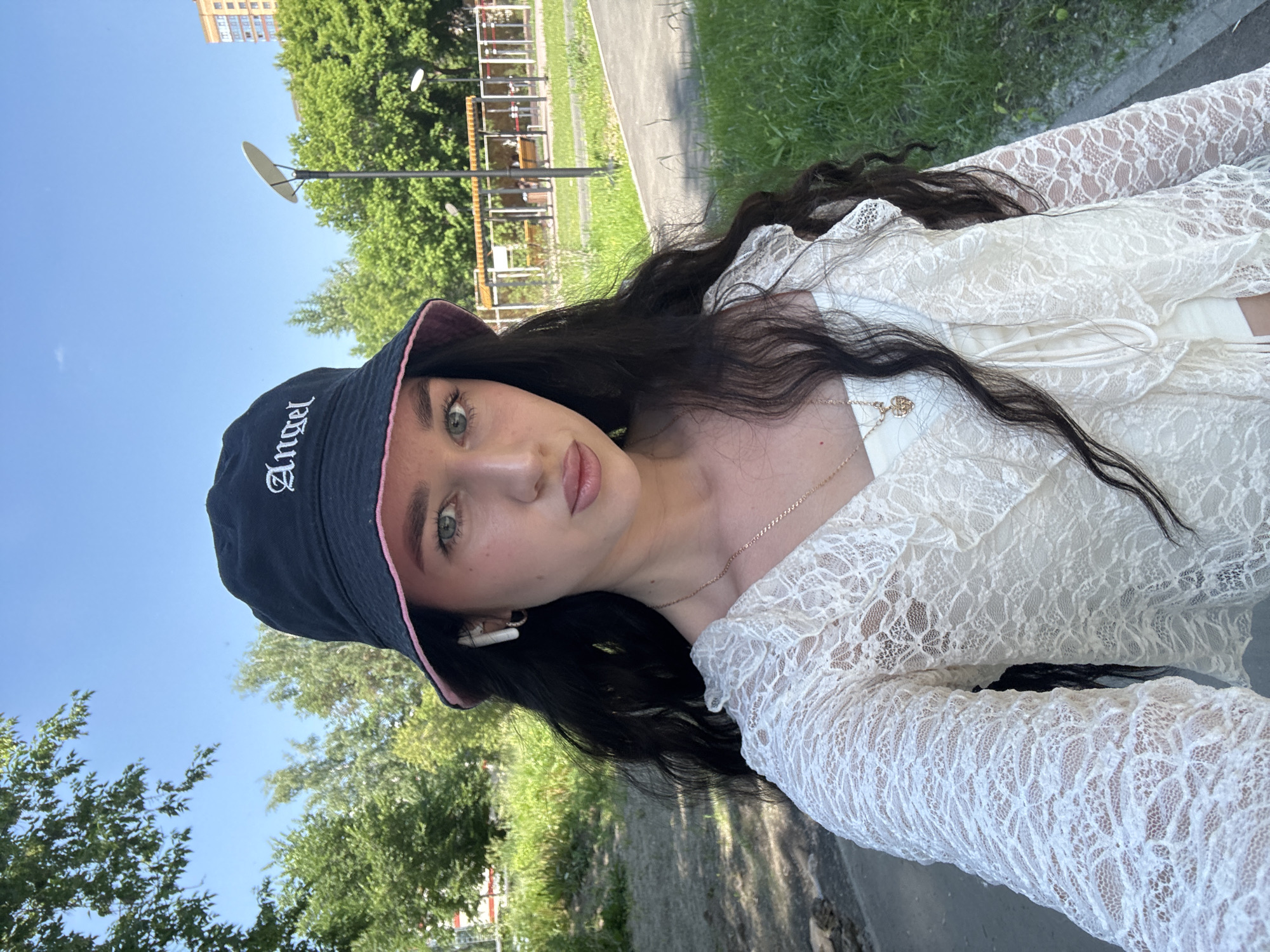 A young woman with long, dark, wavy hair is pictured outdoors, wearing a white lace top and a dark bucket hat with "Angel" embroidered in white gothic script. She has light green eyes and is wearing makeup. She has white wireless earbuds in her ears and a delicate gold necklace with a small pendant. The image is a close-up selfie, taken from a slightly low angle.
The background consists of a blue sky with some clouds, lush green trees, and a paved path, suggesting a park or a residential area on a sunny day. There are also some structures visible in the distance, including a tall building and playground equipment. The overall impression is one of a casual, sunny day spent outdoors. The lighting indicates it is daytime, likely late morning or afternoon. The image appears to have been taken in Novosibirsk, Russia, based on the location context provided.