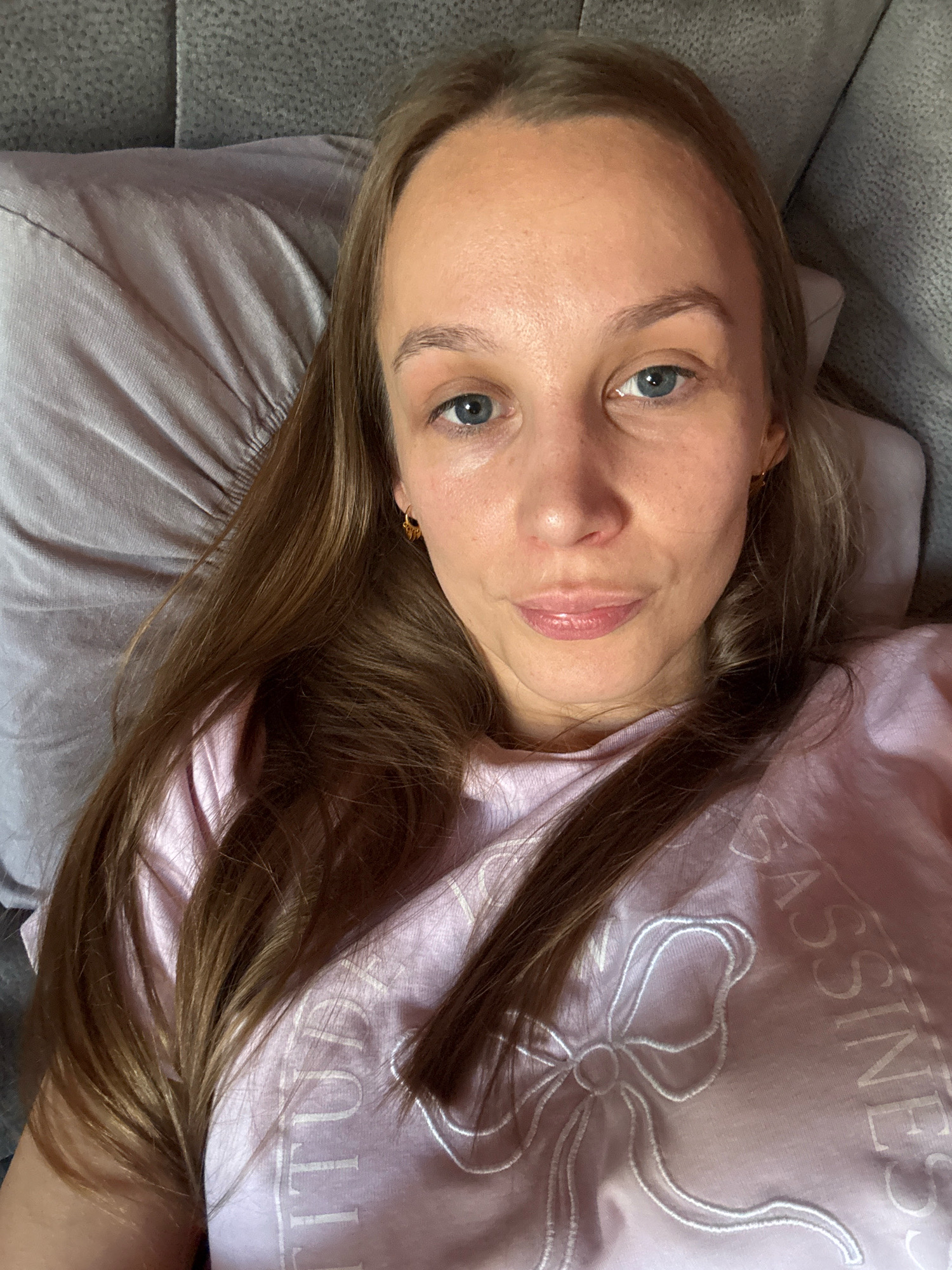 In Überherrn, Germany, a young woman with long, brown hair and blue eyes lies on a couch, looking at the camera. She is wearing a light pink t-shirt with a white bow design and some text. The couch is upholstered in a grey fabric, and a grey pillow is visible behind her head. The lighting is soft and appears to be natural light coming from the side.