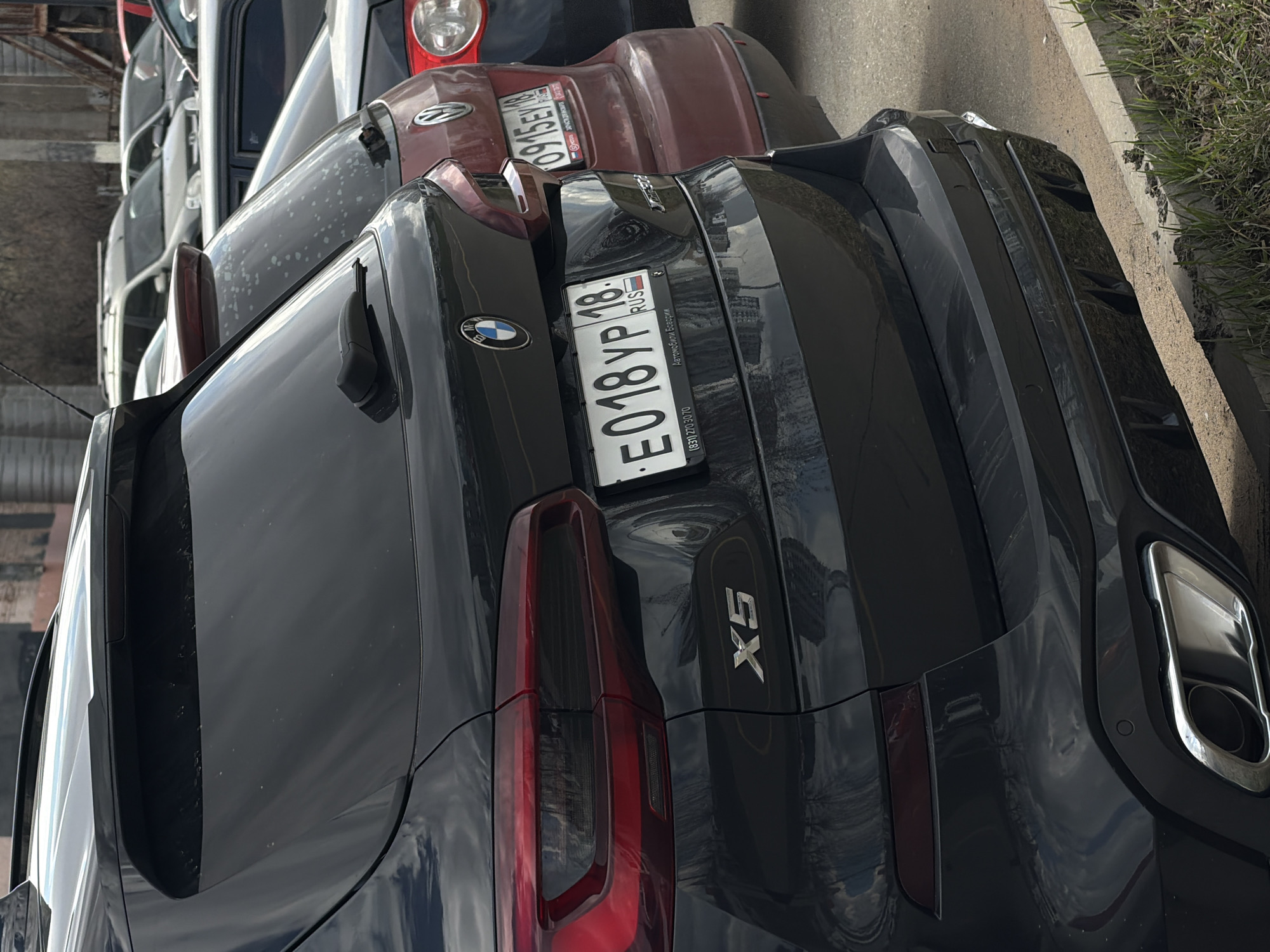 The image shows a black BMW X5 parked on a street. The license plate number is E018YP18, registered in Russia. Other cars are parked in the background. The scene appears to be in an urban area. There is no information to identify specifically the location in the city of Izhevsk.