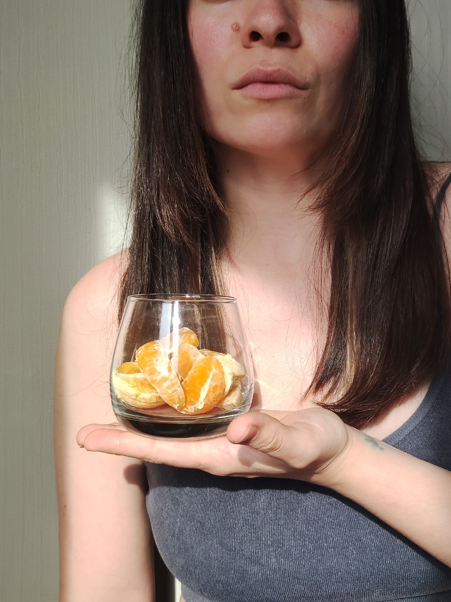 A woman is holding a glass filled with peeled orange slices in her left hand. She is wearing a gray tank top. The background is a light beige wall with a vertical line of light shining on it. The location is Unknown, Unknown.