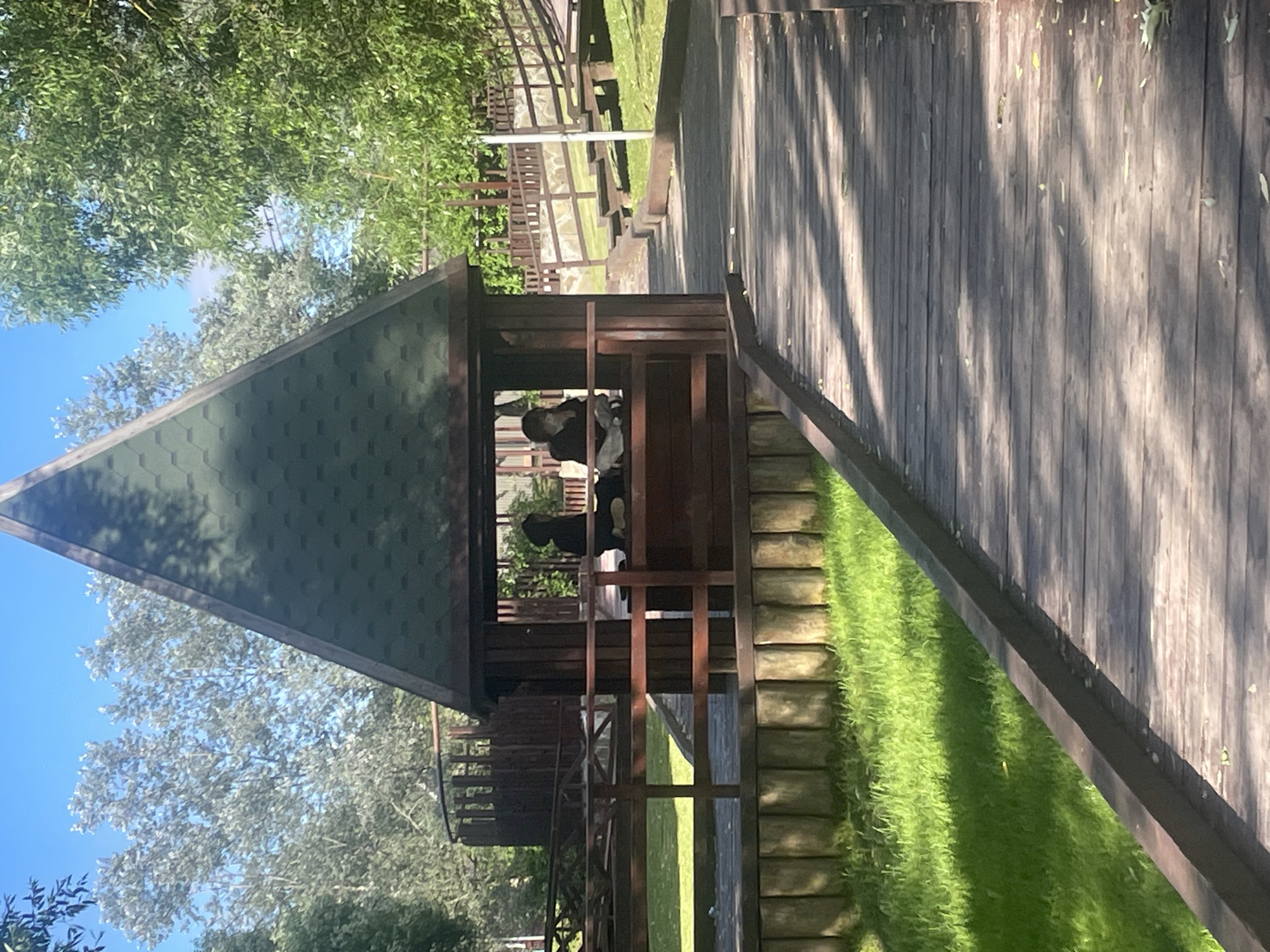 A wooden boardwalk leads through a grassy area with trees and a small wooden gazebo. Two people are seated inside the gazebo. The gazebo has a dark green, hexagonal roof and wooden walls. The boardwalk is made of weathered planks and has a wooden railing along its edge. Sunlight filters through the trees, casting shadows on the boardwalk and grass. The background shows more trees, a wooden fence, and a staircase. This scene is located in Marushkino, Russia.