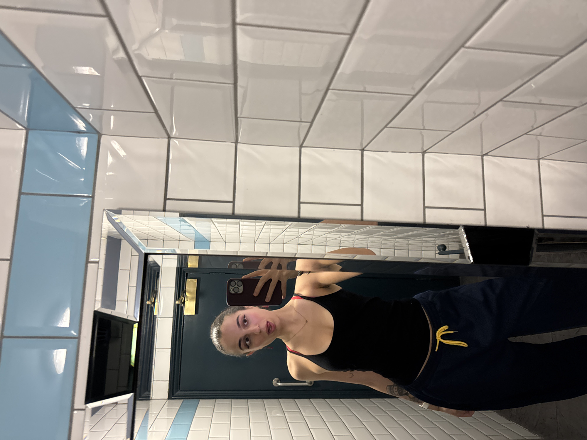 A person is taking a selfie in a mirrored bathroom in what appears to be a public restroom. The background has a blue and white tile pattern. The person is wearing a black tank top and blue sweatpants with a yellow drawstring. A door with gold hardware is in the background. The subway style tiles and door fixtures could possibly place this in London, United Kingdom.
