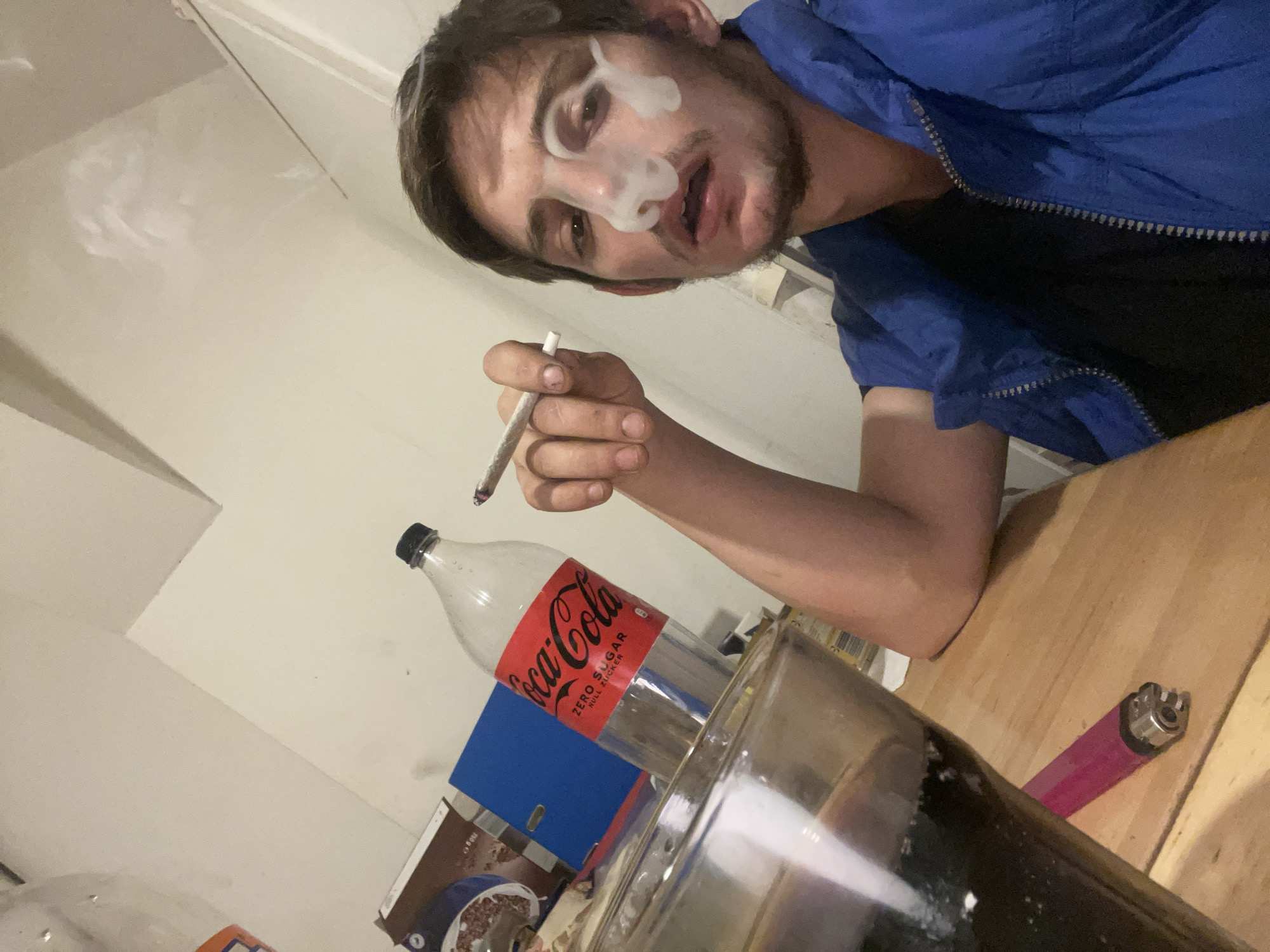 The image shows a person with smoke emanating from their nose and mouth, holding a lit cigarette in their right hand. They are wearing a blue jacket over a dark top. On the table in front of the person are a Coca-Cola Zero Sugar bottle, a glass filled with a dark liquid, a pink lighter, and a box of cereal. The environment appears to be indoors, possibly a residence, with white walls. The location is listed as Unknown, Unknown.