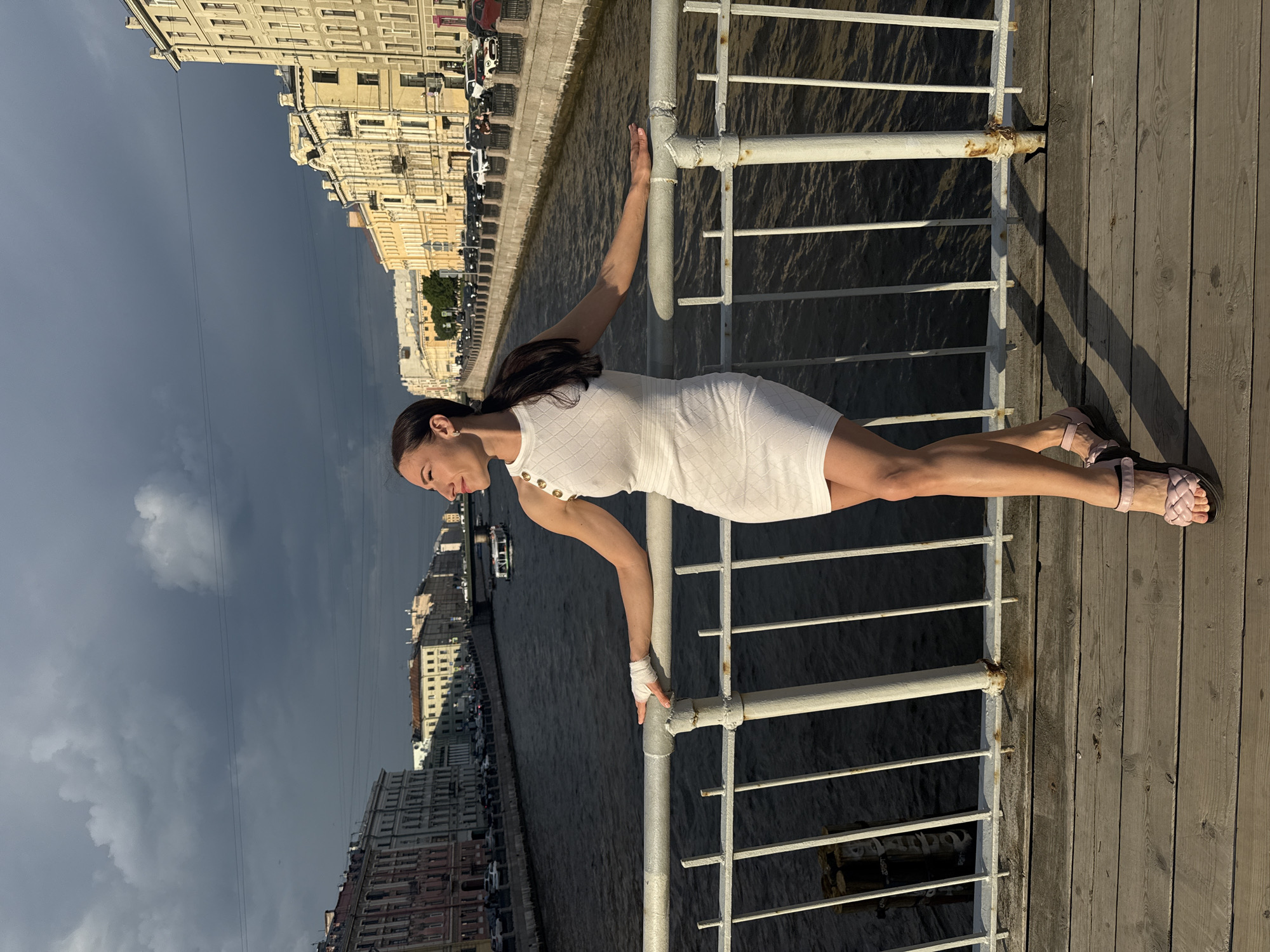 A woman in a white, textured short-sleeved dress with gold buttons is captured in a dynamic pose on a wooden pier, appearing to hold herself horizontally by gripping a white metal railing with both hands. Her dark hair is pulled back, and she wears light pink sandals. A bandage or wrap is visible on her right wrist. She looks focused, slightly downwards.
The setting is a waterfront area, likely a promenade or pier, in Saint Petersburg, Russia. Behind the railing, a calm body of water, resembling a river or canal, stretches into the distance. The opposite bank is lined with classical European-style buildings, characteristic of Saint Petersburg architecture, with some cars visible along the street. A small boat can be seen further down the waterway.
The time of day appears to be late afternoon or early evening, indicated by the long shadows cast by the railing and the woman onto the wooden planks of the pier. The sky presents a dramatic contrast, with dark grey, ominous clouds dominating the upper left portion, while the sky becomes lighter towards the right, suggesting shifting weather patterns or an approaching storm. No visible text is present in the image.