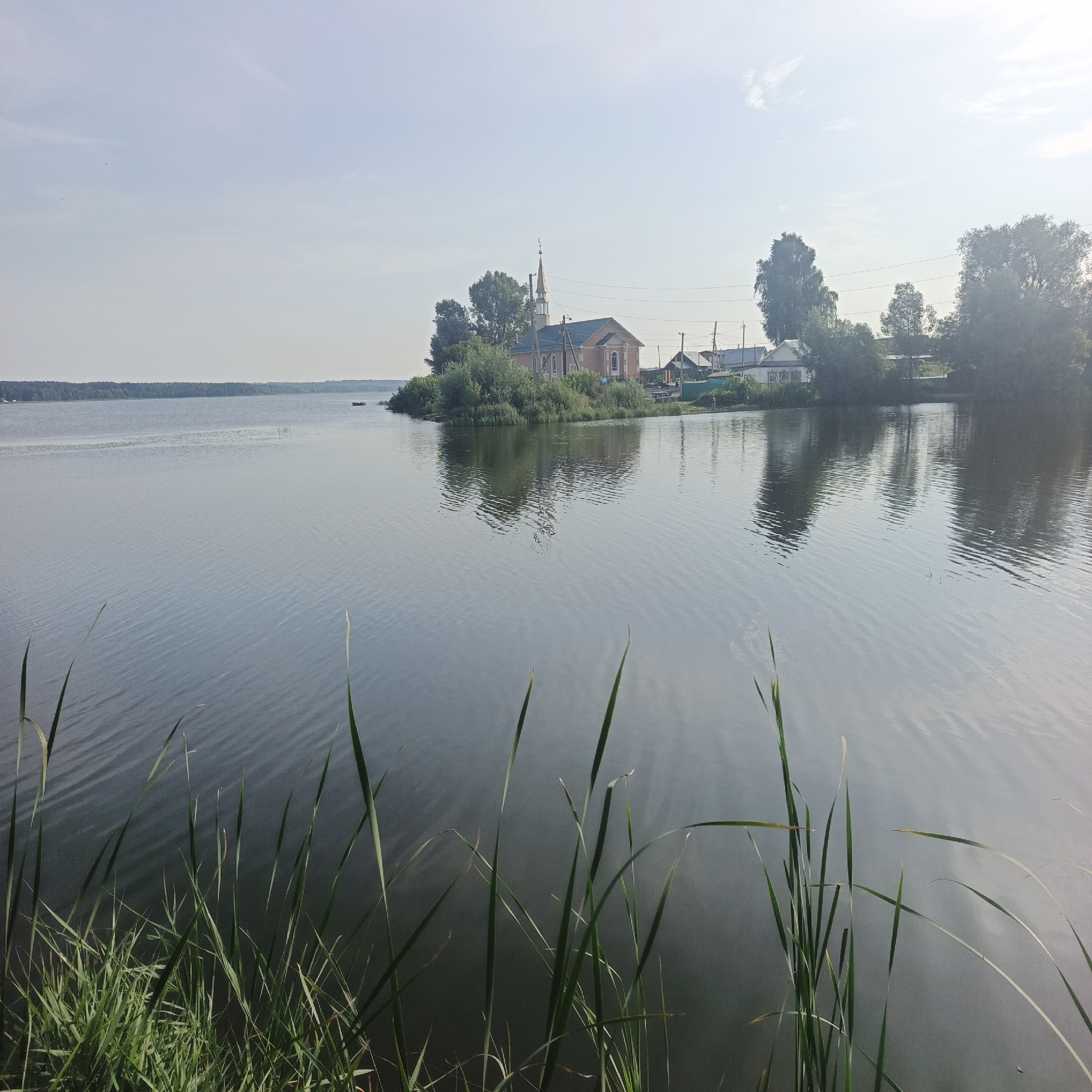 A wide shot captures a serene lakeside scene in Kambarka, Russia, under a pale, hazy sky. In the mid-ground, a small, verdant island juts into the calm, rippling water, adorned with a red-brick building featuring a golden spire topped with a crescent moon, suggesting it is a mosque or religious structure. Dense green foliage surrounds the building, and several trees are scattered across the island. Beyond the island, a few simple houses and power lines are visible, hinting at a nearby settlement. The foreground is dominated by the dark, reflective surface of the lake, with tall green reeds and grasses growing along the water's edge. The overall atmosphere is tranquil and slightly overcast, with the reflection of the island and the sky subtly visible on the water's surface.