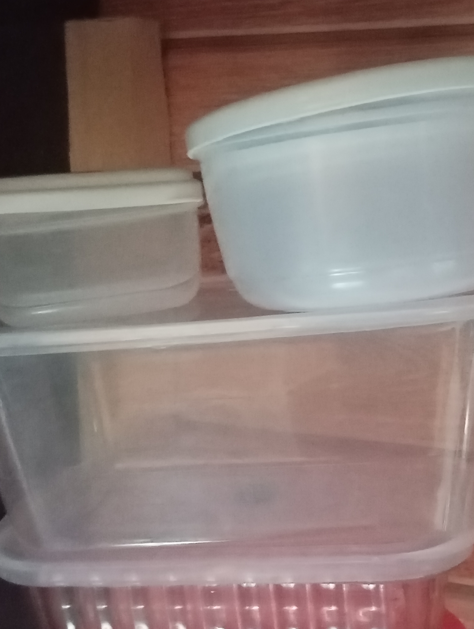 A clear plastic storage container with a red ribbed bottom and a clear lid is the primary subject. It is stacked on top of another clear plastic storage container with a white lid. A third, smaller, clear plastic storage container with a white lid is placed on top of the second container. The containers are stacked on a wooden surface. The background is out of focus and appears to be a wooden wall. The image is taken from a medium-high angle.